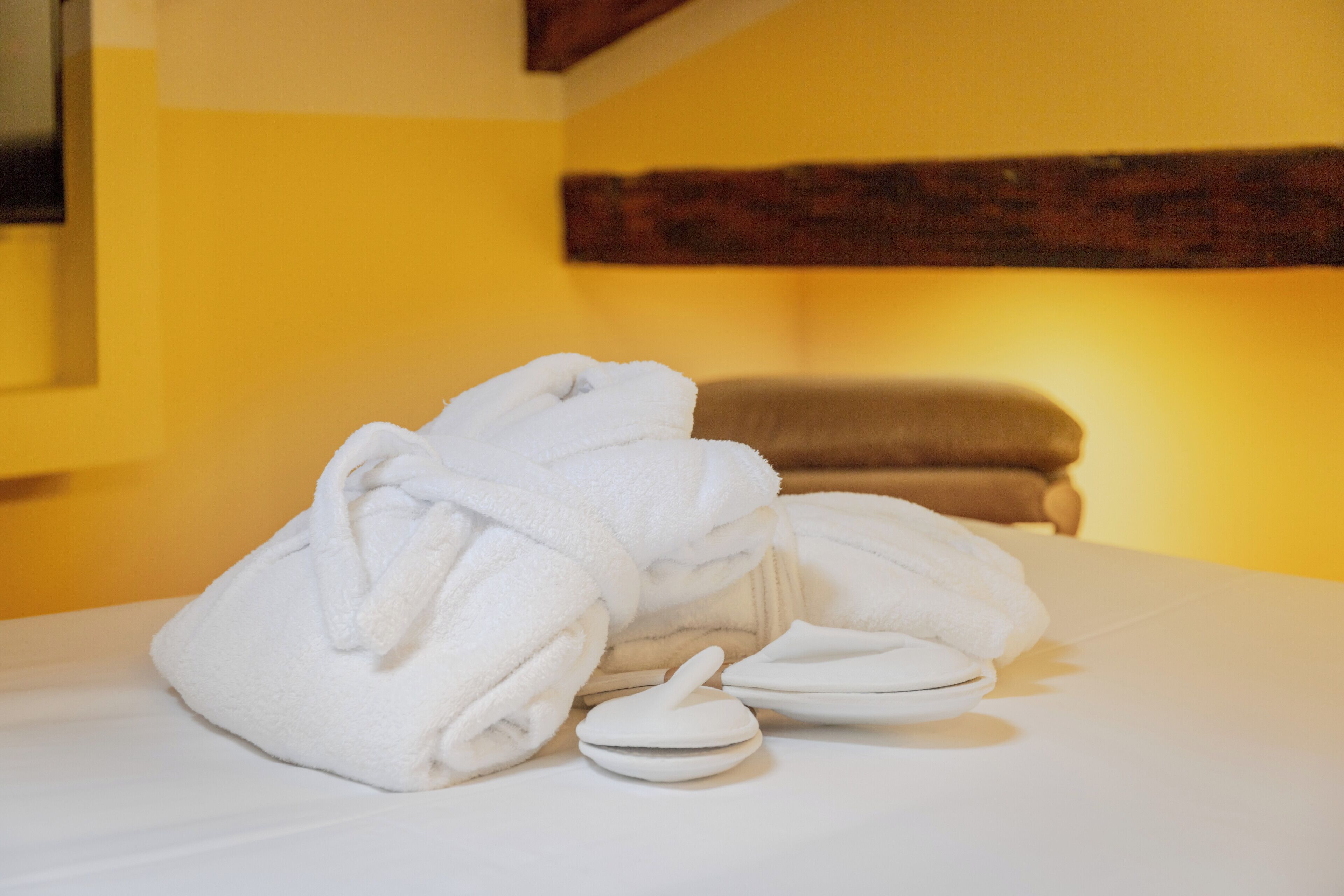 A neatly folded white towel and a white bath mat on a bed in a room with yellow walls and a wooden beam.