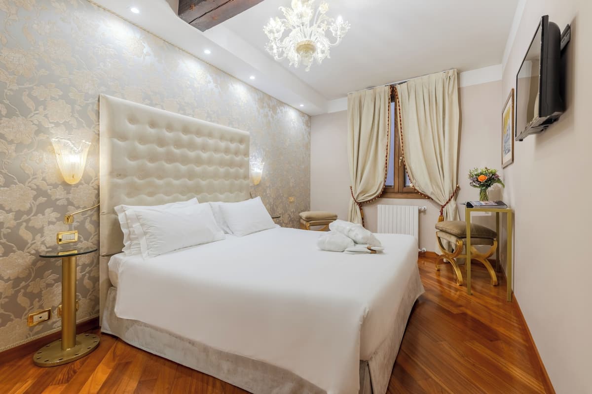 A hotel room with a large bed, a wall-mounted TV, a chandelier, and a small side table.