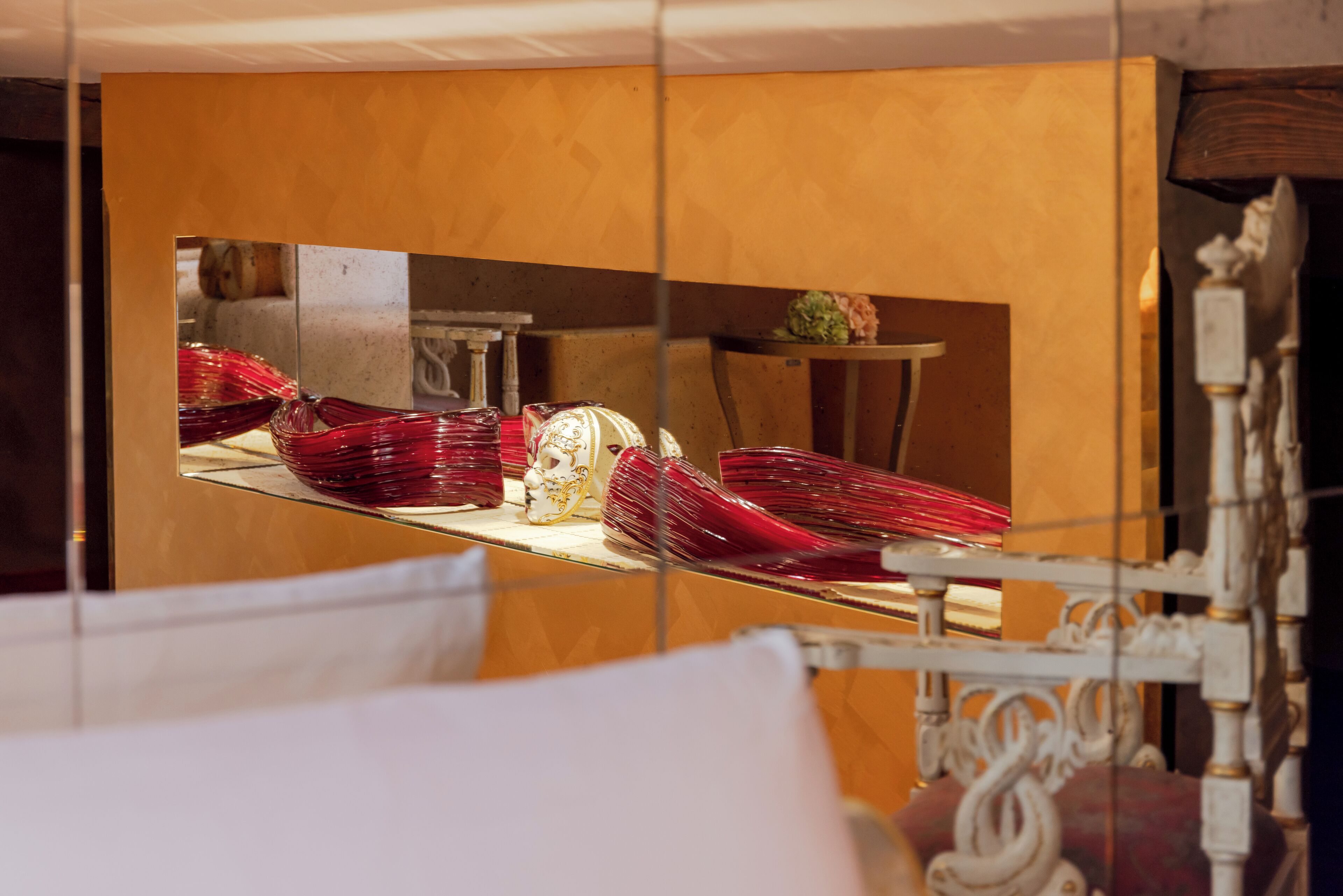 A room with a bed, a mirror reflecting a dining area with red glassware and a white table.