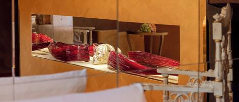 A room with a bed, a mirror reflecting a dining area with red glassware and a white table.