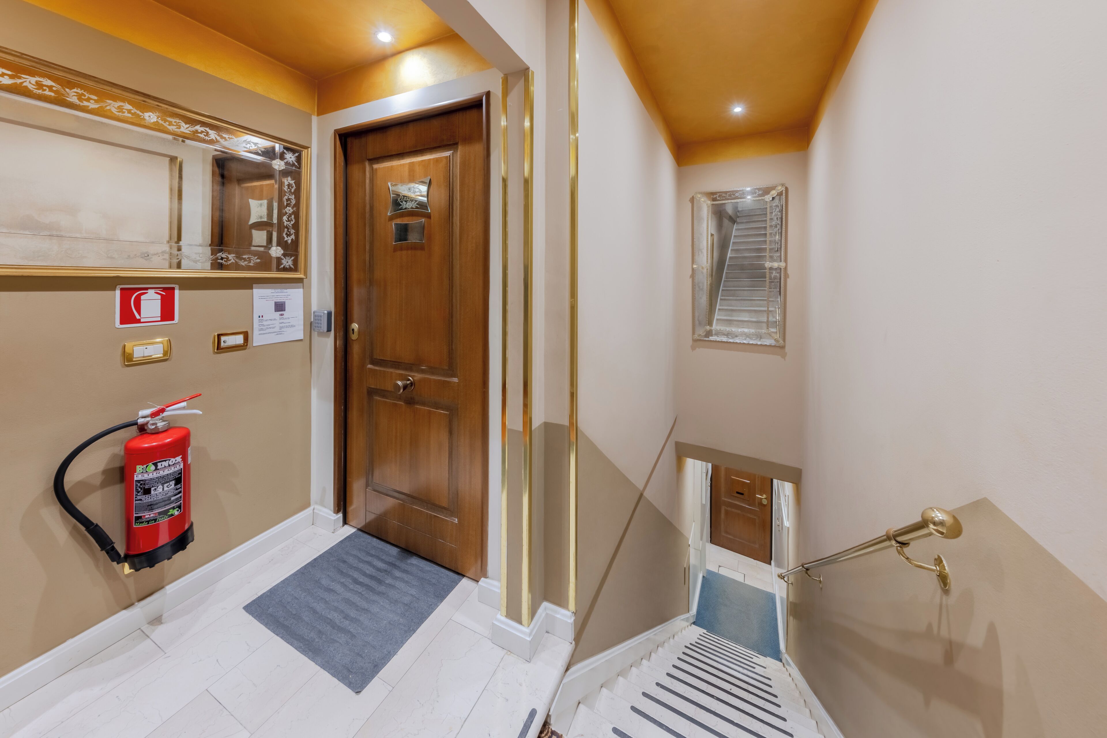 A hotel hallway with a fire extinguisher, a wooden door, a mirror, a window, and a staircase.