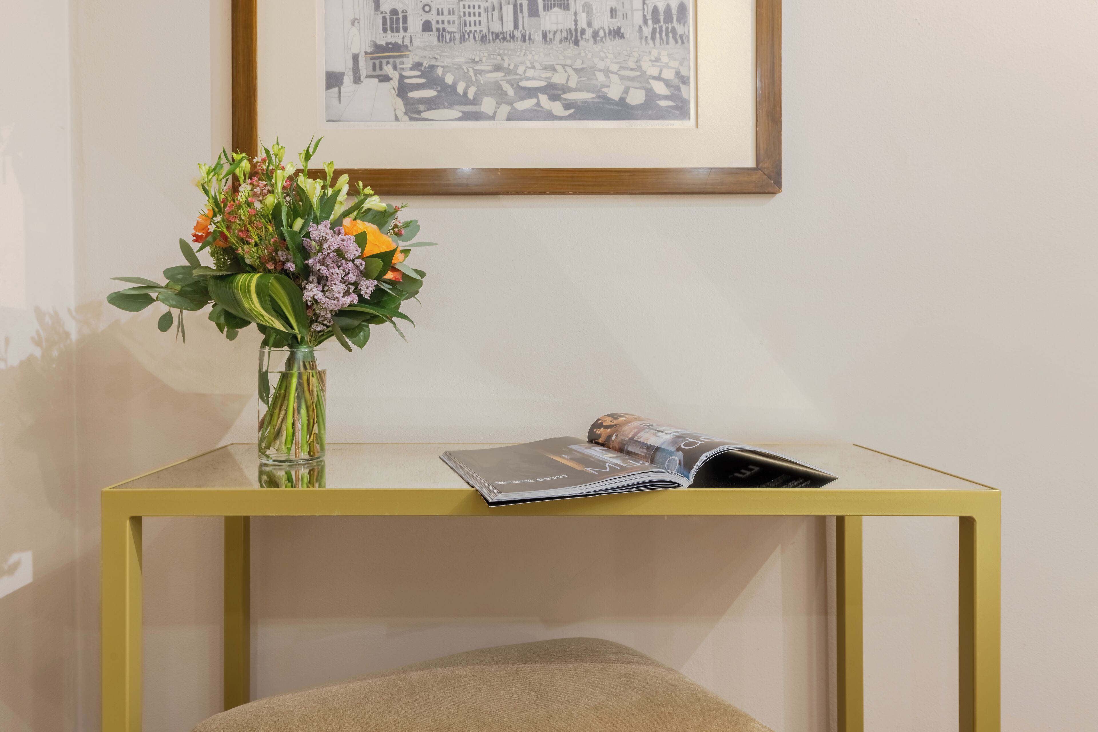 A room with a glass-top table, a framed picture on the wall, and a vase with flowers.