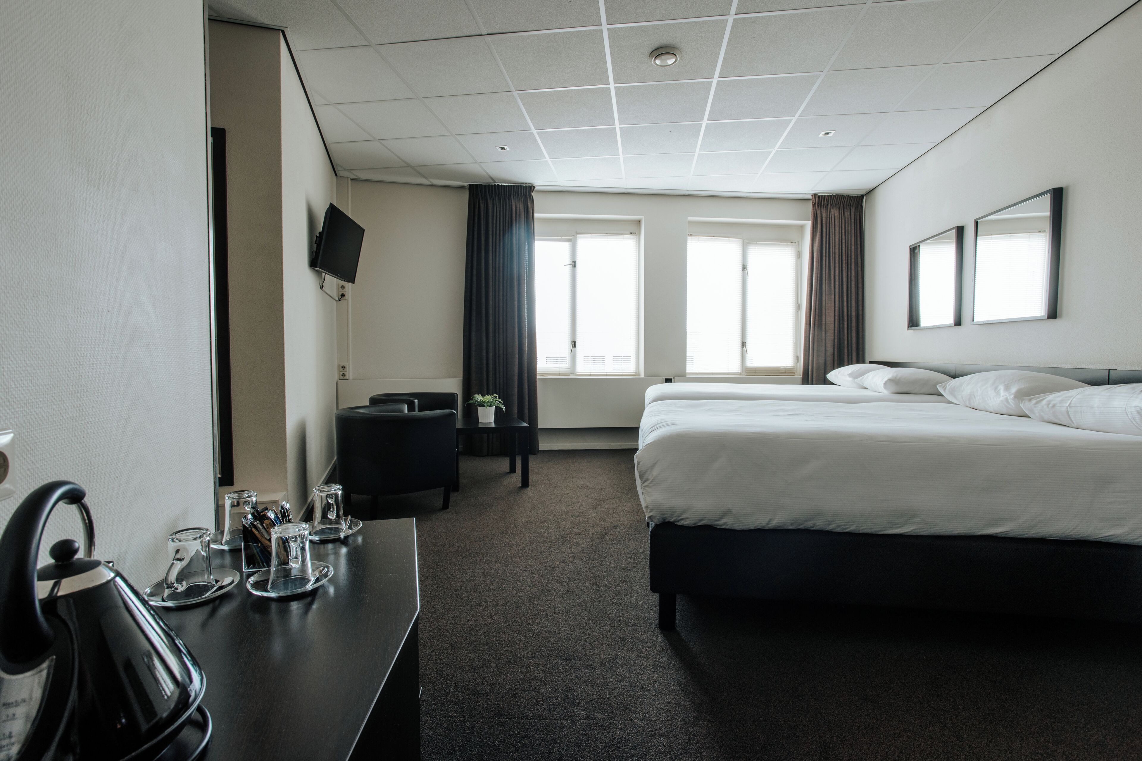 Habitación de hotel con cama, escritorio con hervidor, televisión y ventana con cortinas.