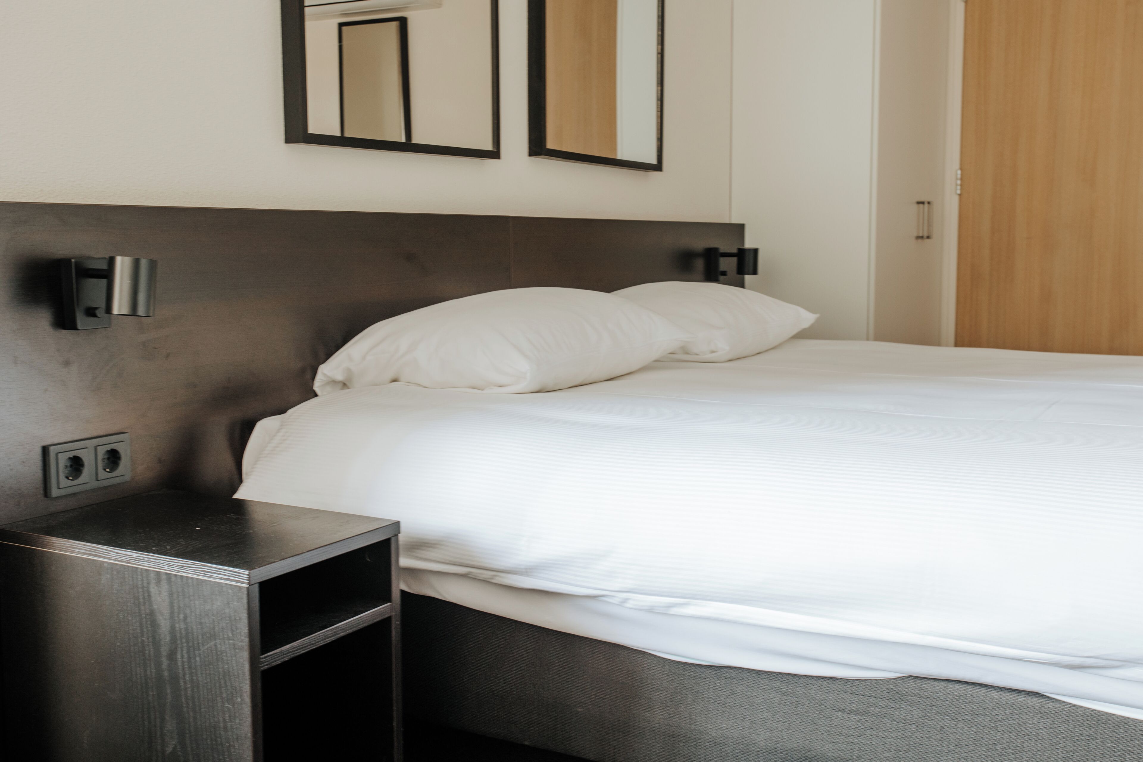 Cama bem arrumada com lençóis brancos e cabeceira escura. Duas luminárias fixadas na parede, uma de cada lado da cama.