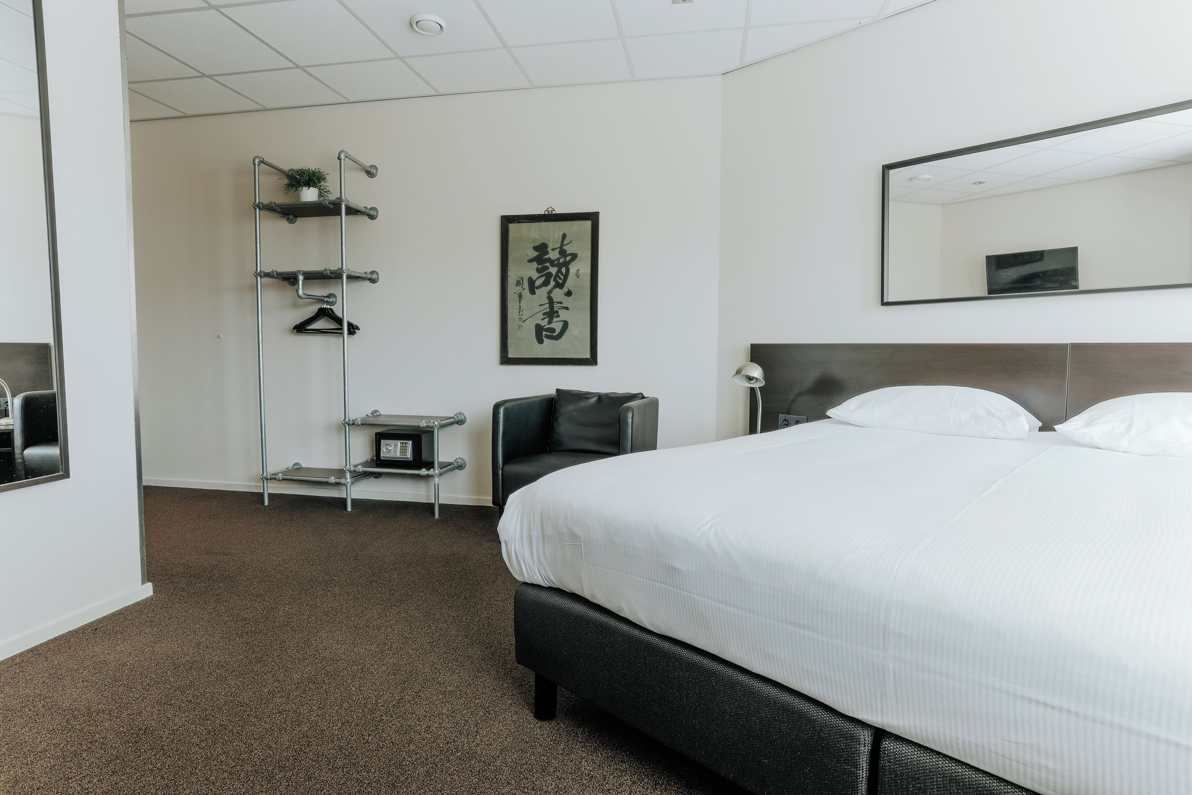 A hotel room with a large bed, a black chair, a desk with a lamp, a mirror, and a wall-mounted shelf with a plant.