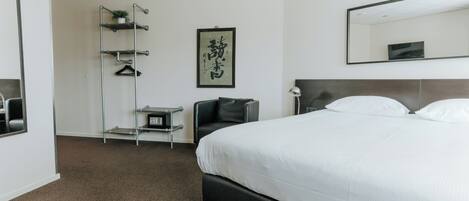 A hotel room with a large bed, a black chair, a desk with a lamp, a mirror, and a wall-mounted shelf with a plant.