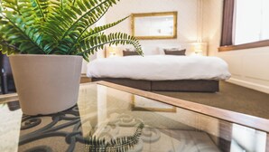 Een hotelkamer met een bed, nachtlampen, een potplant varens en een tafel met glazen blad.