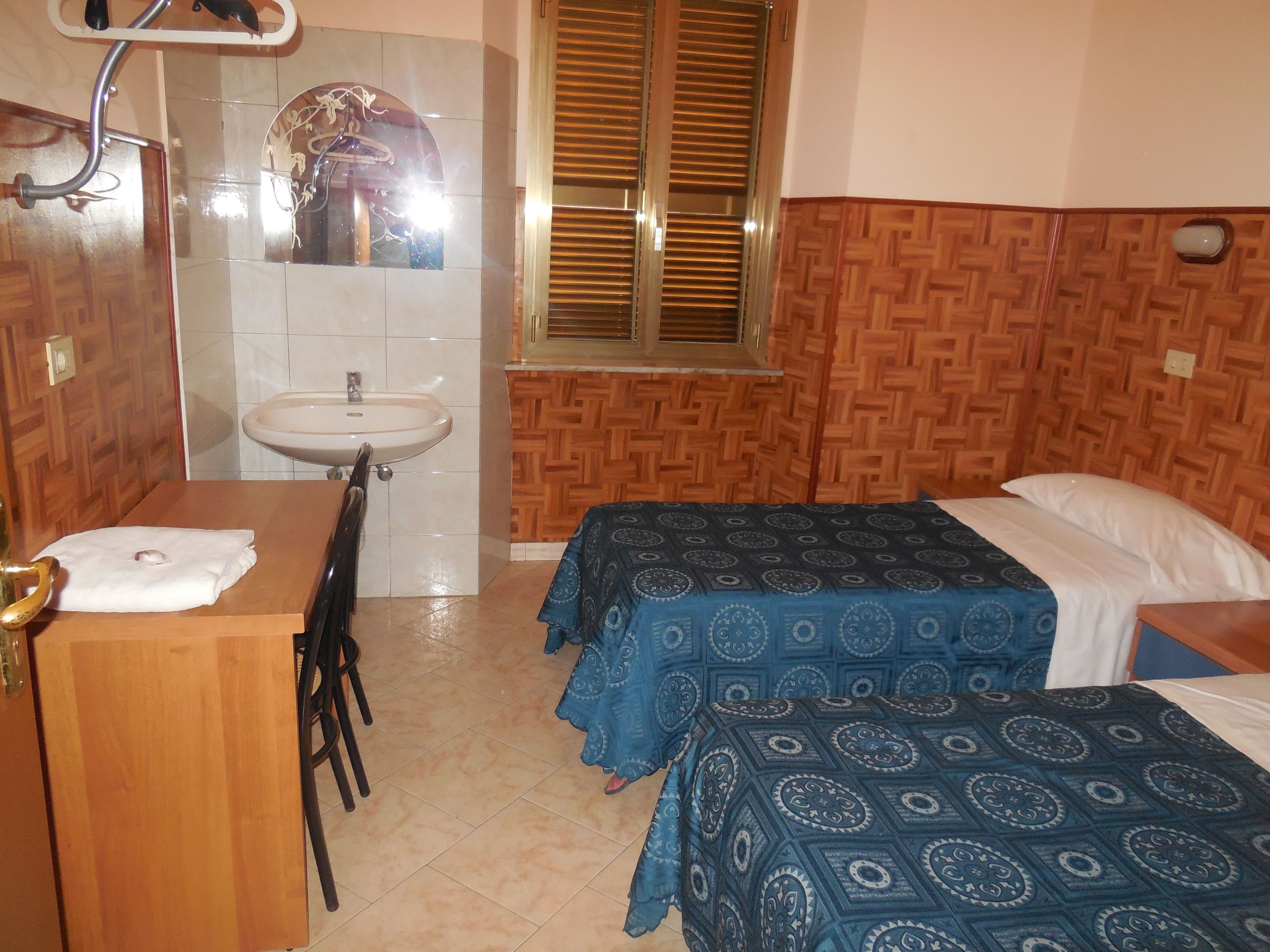 A room with a wooden desk, a sink, a mirror, and two beds with patterned bedspreads.