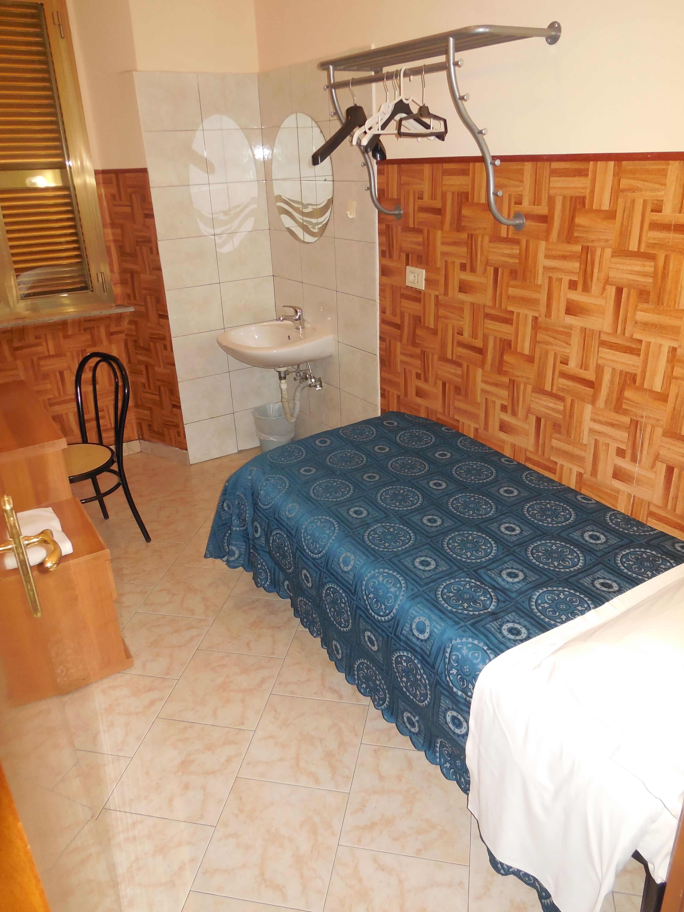 Una habitación pequeña y ordenada con cama, una silla, escritorio y lavamanos.