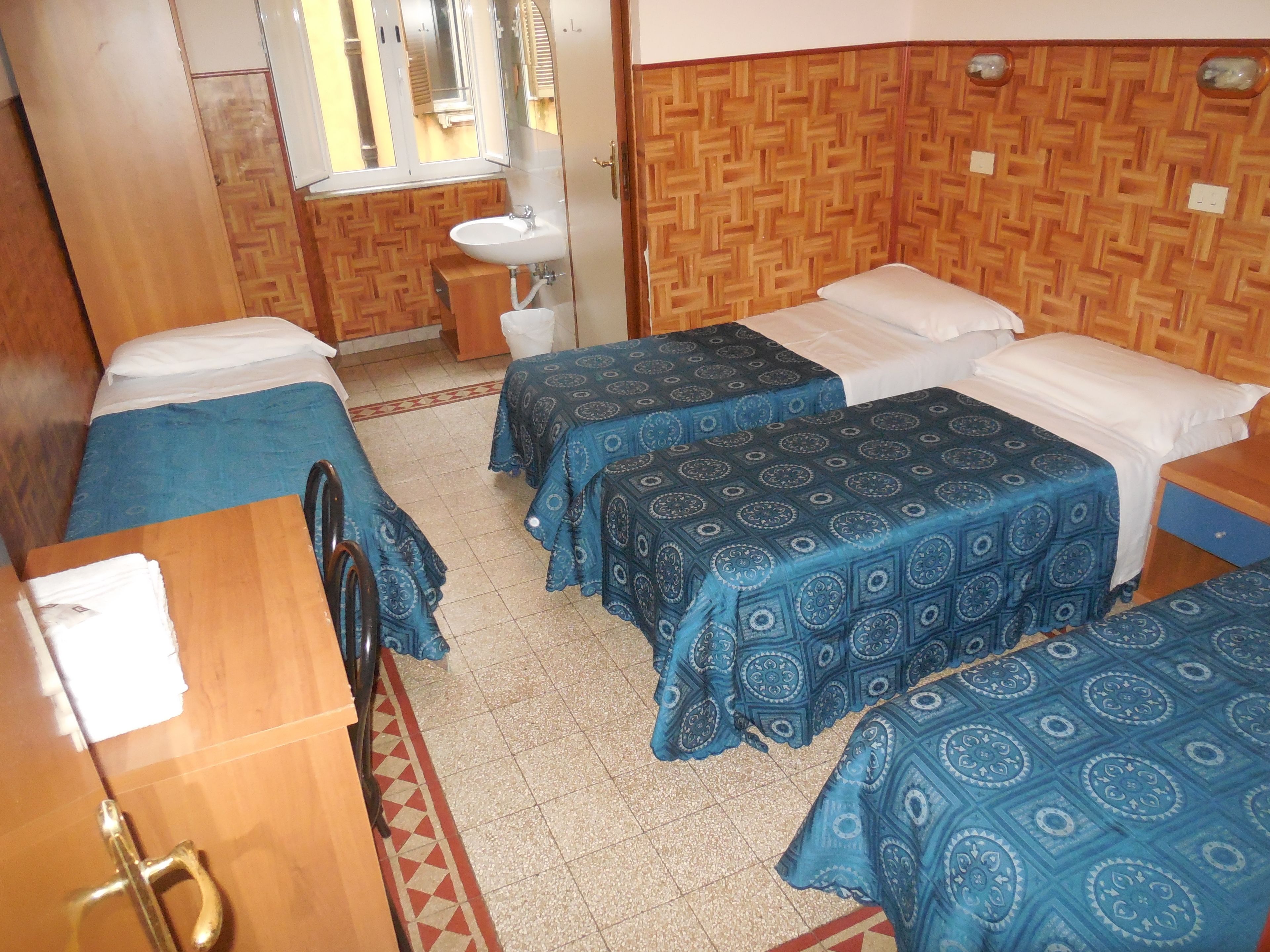 A room with two beds, a wooden wardrobe, a small sink, and a window.