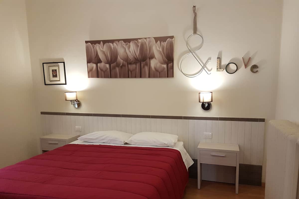 Quarto com colcha vermelha, duas mesas de cabeceira, luminária na parede, quadro emoldurado e decoração de parede com a inscrição 'Love &'.