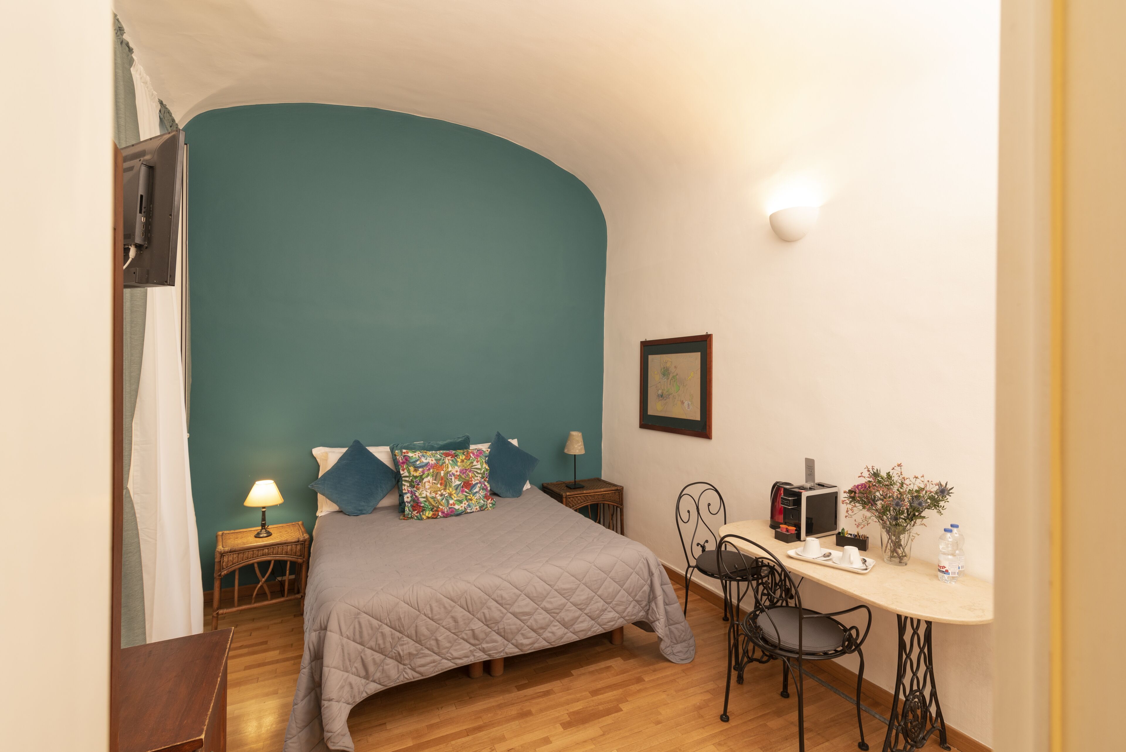 A bedroom with a bed, a desk, a chair, a lamp, and a small table with a vase and a bottle.