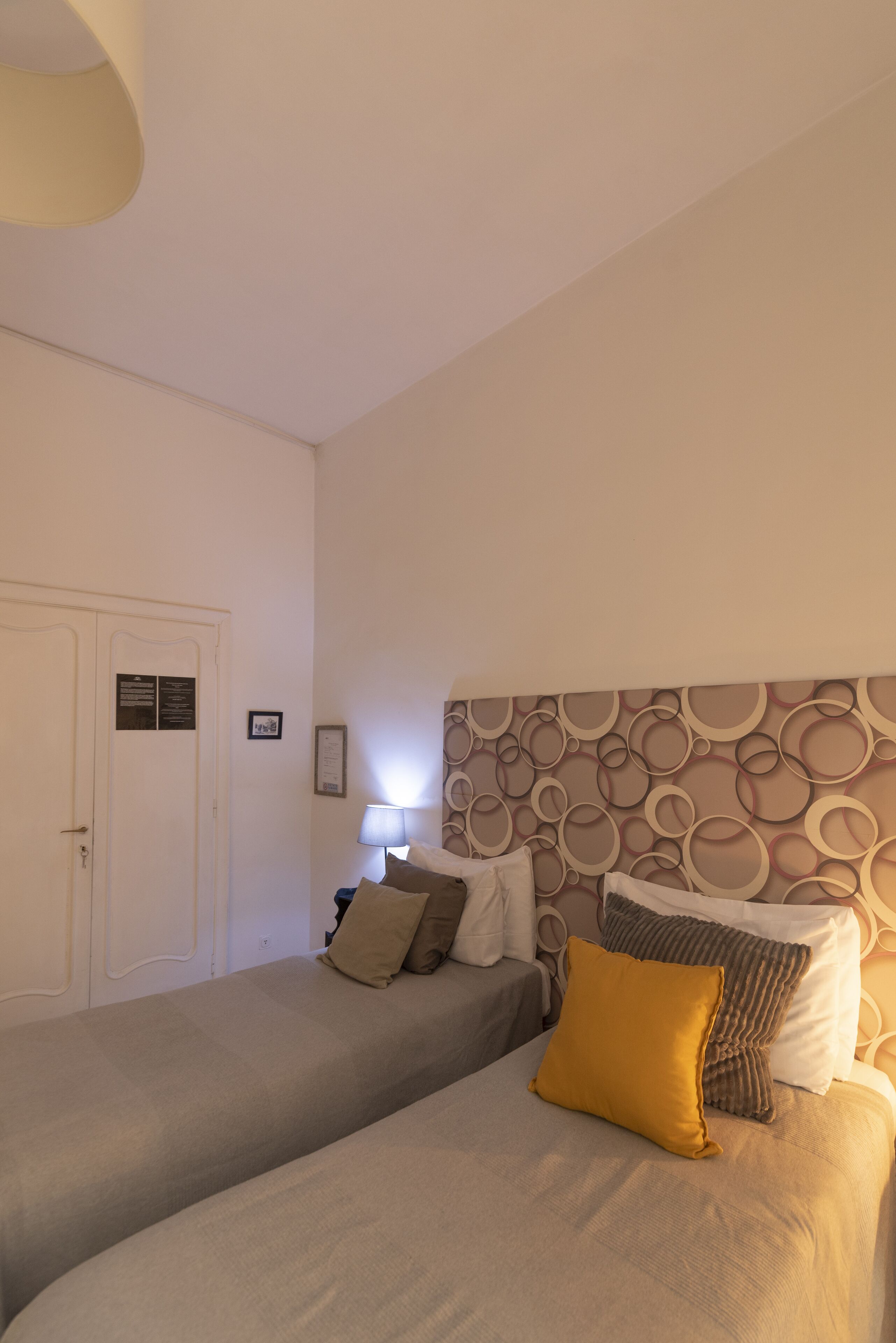 Habitación de hotel con dos camas, cabecera con diseños circulares, una puerta y una lámpara fijada a la pared.