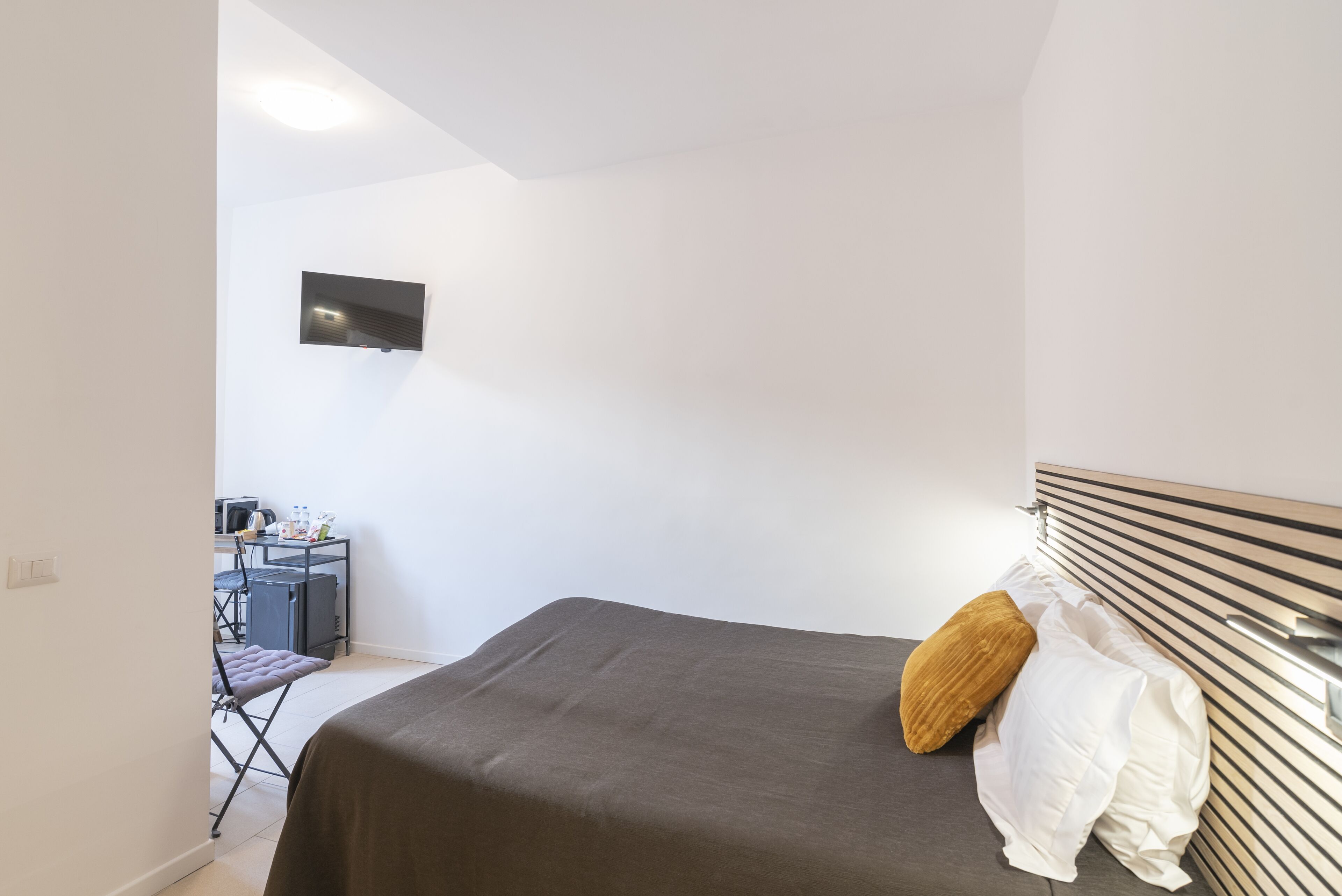 A bedroom with a bed, a TV mounted on the wall, a desk with a chair, and a window.