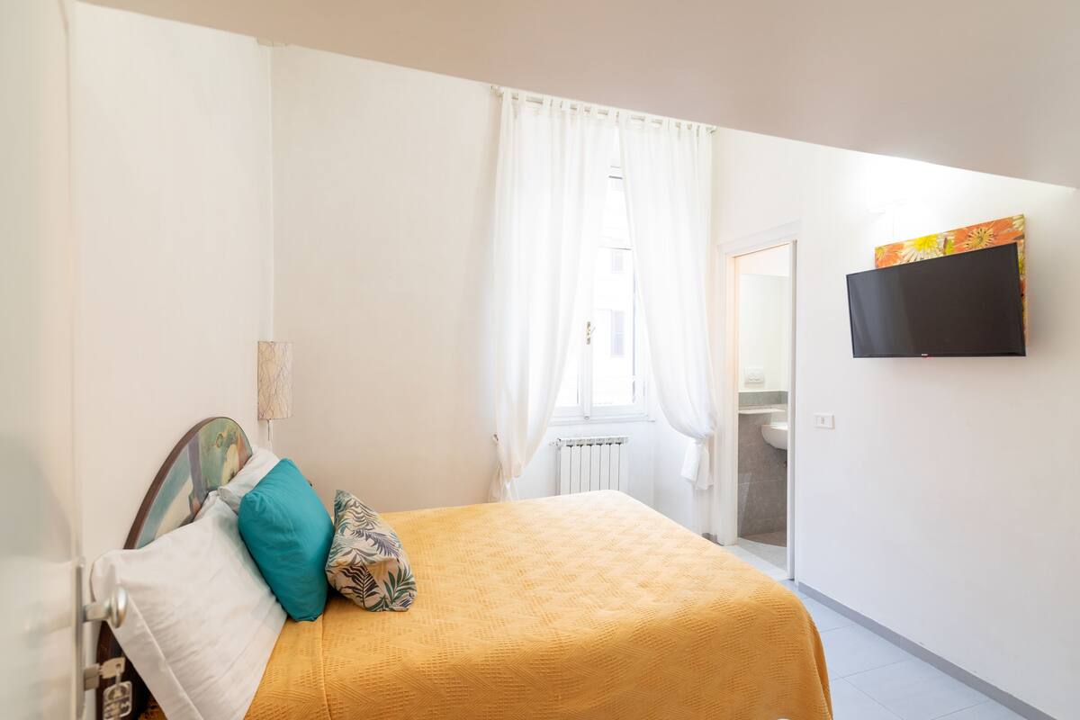 A bedroom with a bed, white curtains, a television on the wall, and a bathroom visible through an open door.