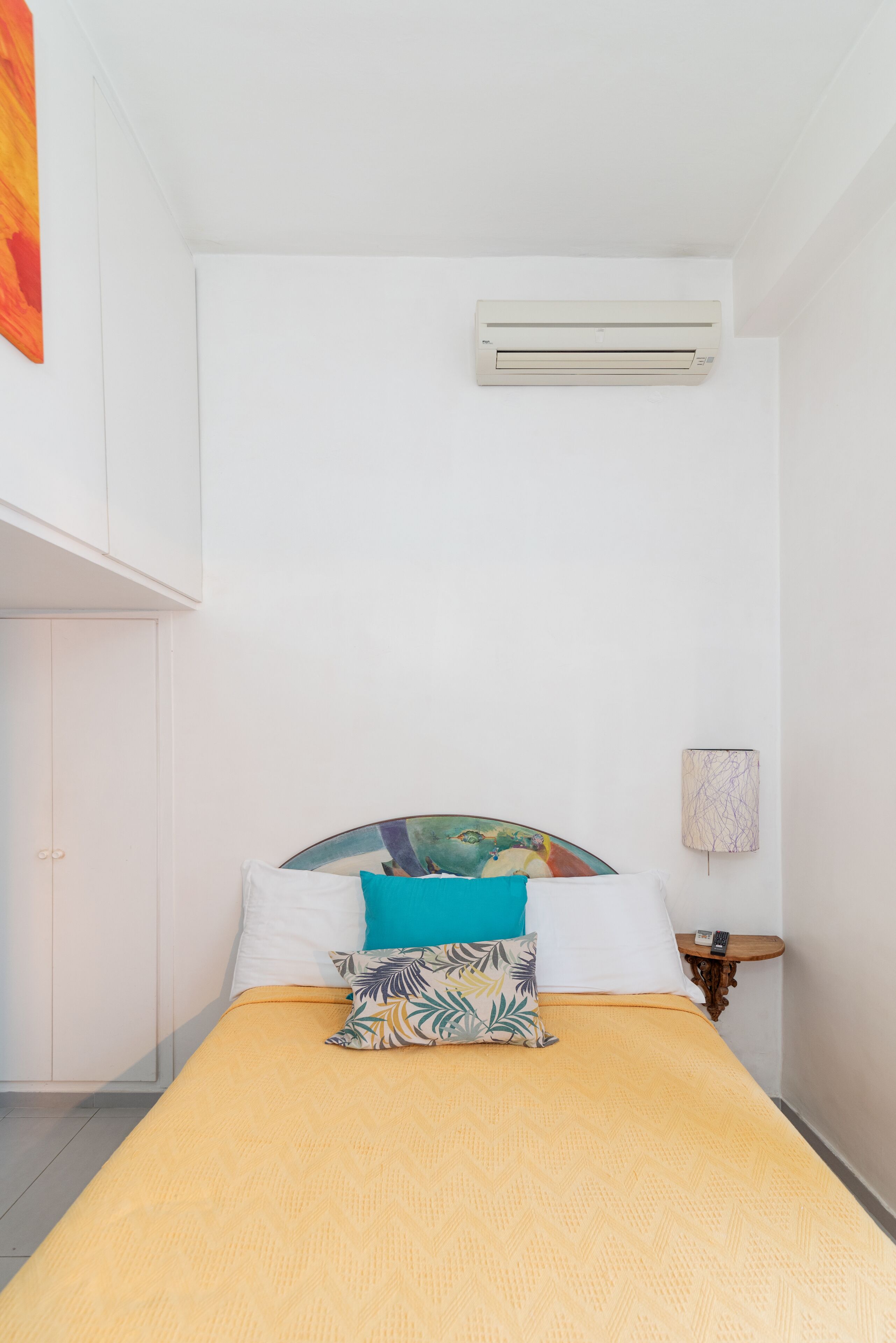 A bed with a yellow bedspread, white pillows, and a colorful headboard. There is a wall-mounted air conditioner above the bed and a small table with a lamp beside it.