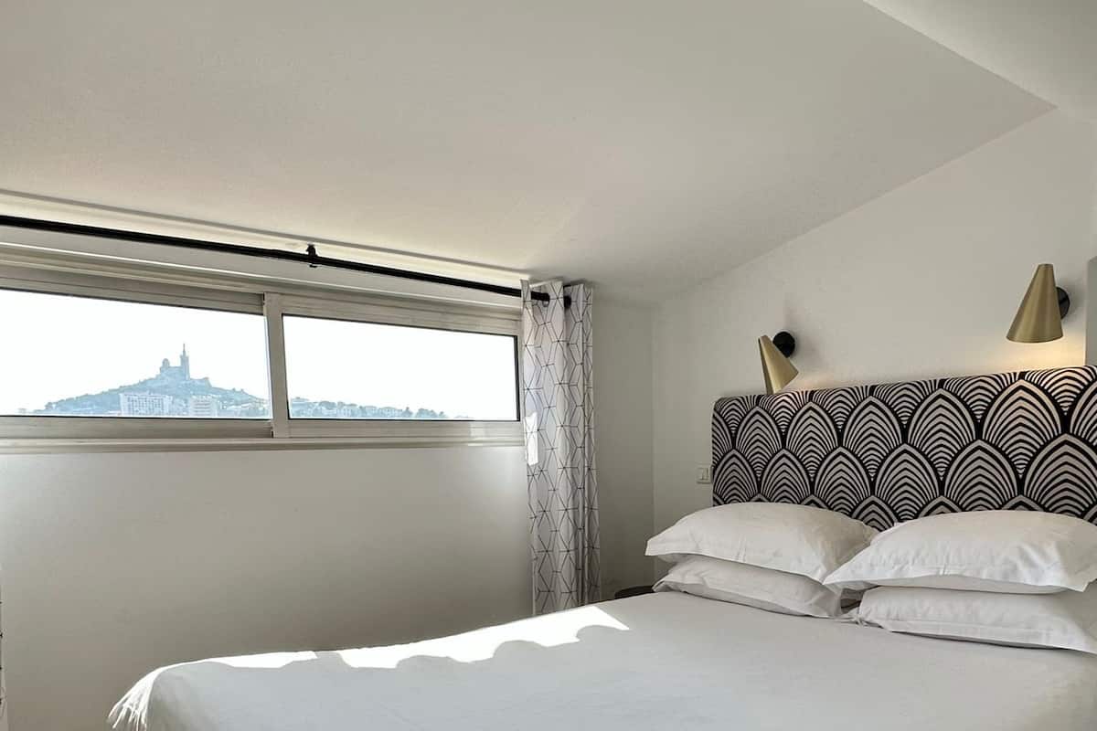 Une chambre à coucher avec un grand lit, une fenêtre avec des rideaux, une tête de lit à motifs et une vue sur un bâtiment situé au loin.