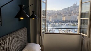 Un dormitorio con cama, vista de un paisaje urbano a través de un ventanal grande y una lámpara fijada en la pared.