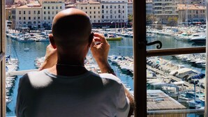 Un hombre mirando por una ventana hacia un puerto deportivo con barcos y edificios al fondo.