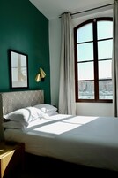 A bedroom with a bed, a framed picture on the wall, a window with curtains, and a view of buildings outside.