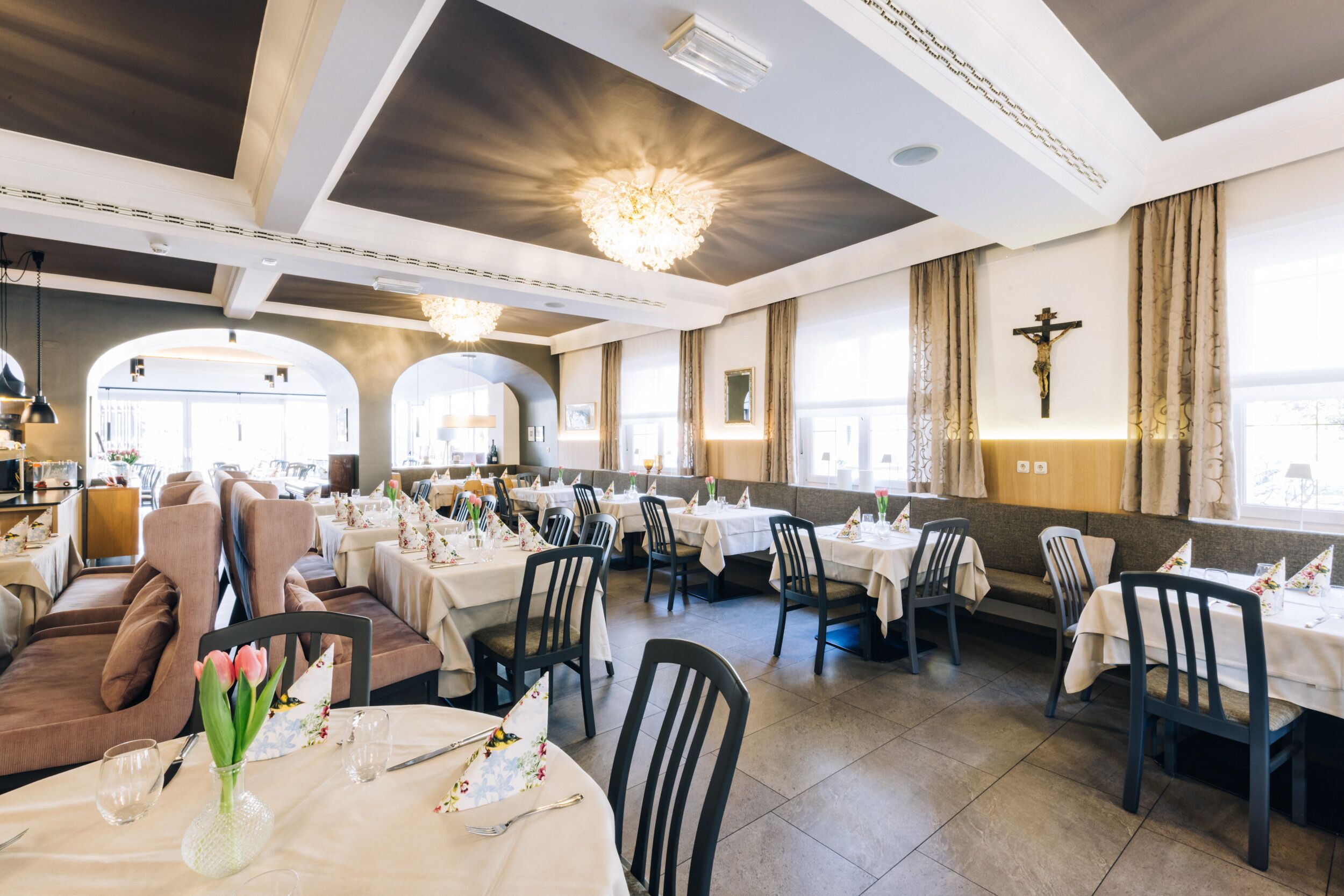 Um restaurante com várias mesas e cadeiras, um lustre e uma cruz na parede.