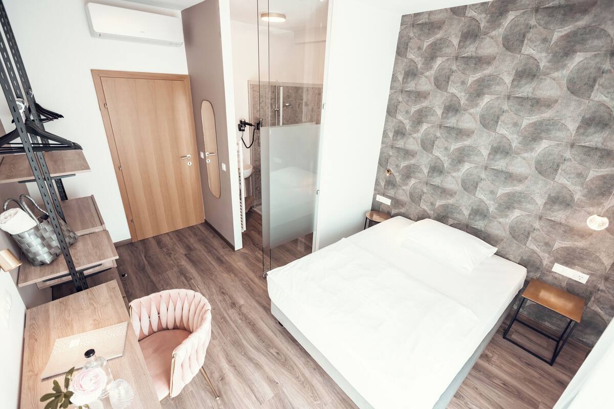 A modern bedroom with a bed, a wooden floor, a stone wall, a glass shower, and a wooden door.