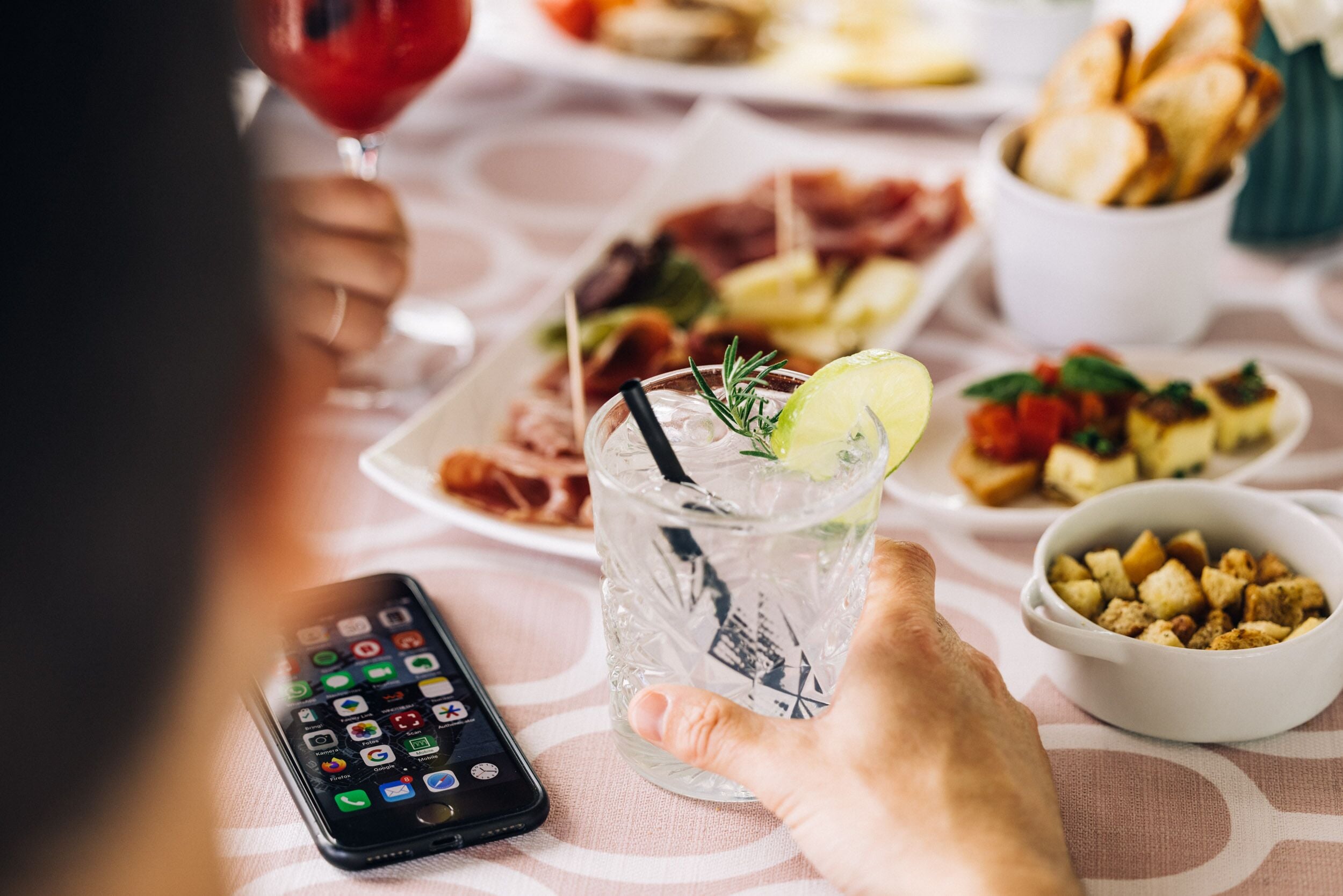 Une personne tenant un verre avec une paille et une tranche de citron vert, un smartphone posé sur une table, et divers aliments, comme du pain et un bol de croûtons.