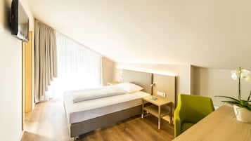 A hotel room with a bed, a desk, a chair, a TV mounted on the wall, and a potted plant.