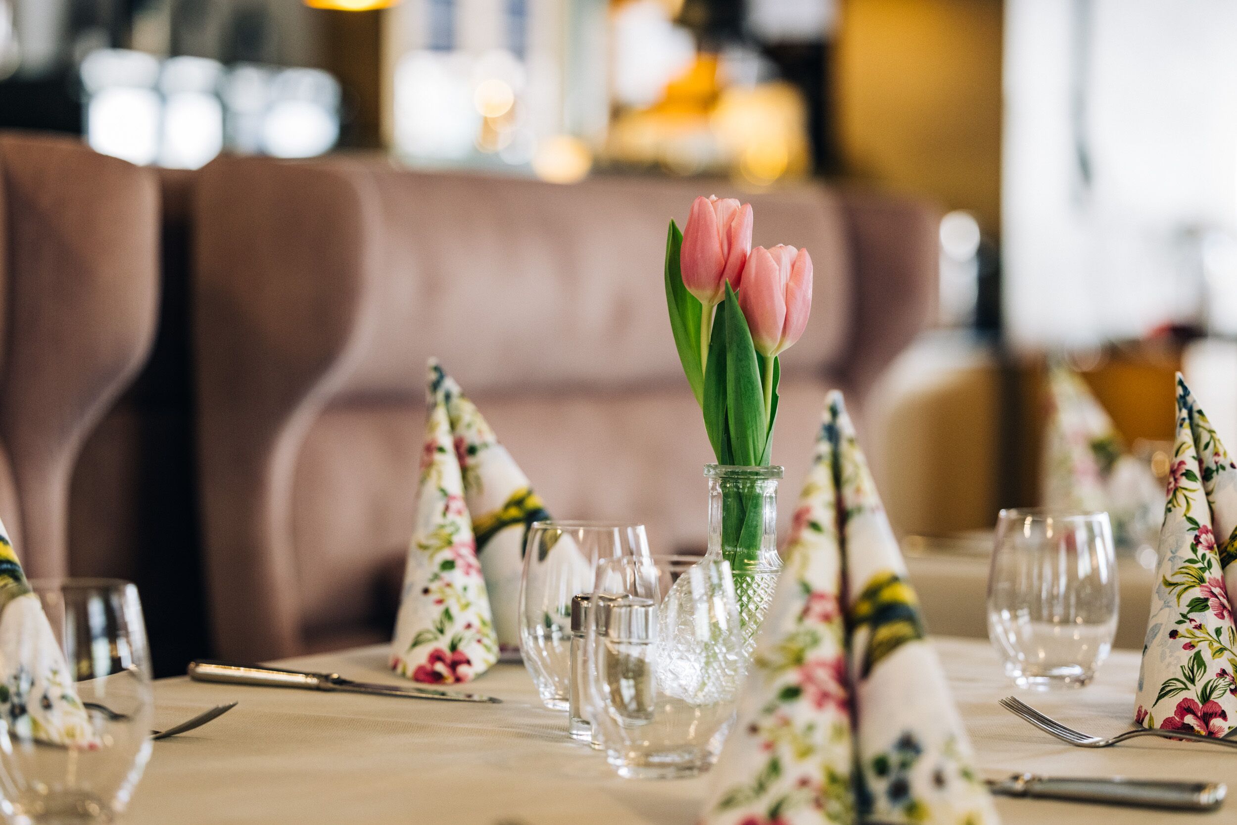 A dining table set with clear glassware, a vase with pink tulips, and floral napkins.