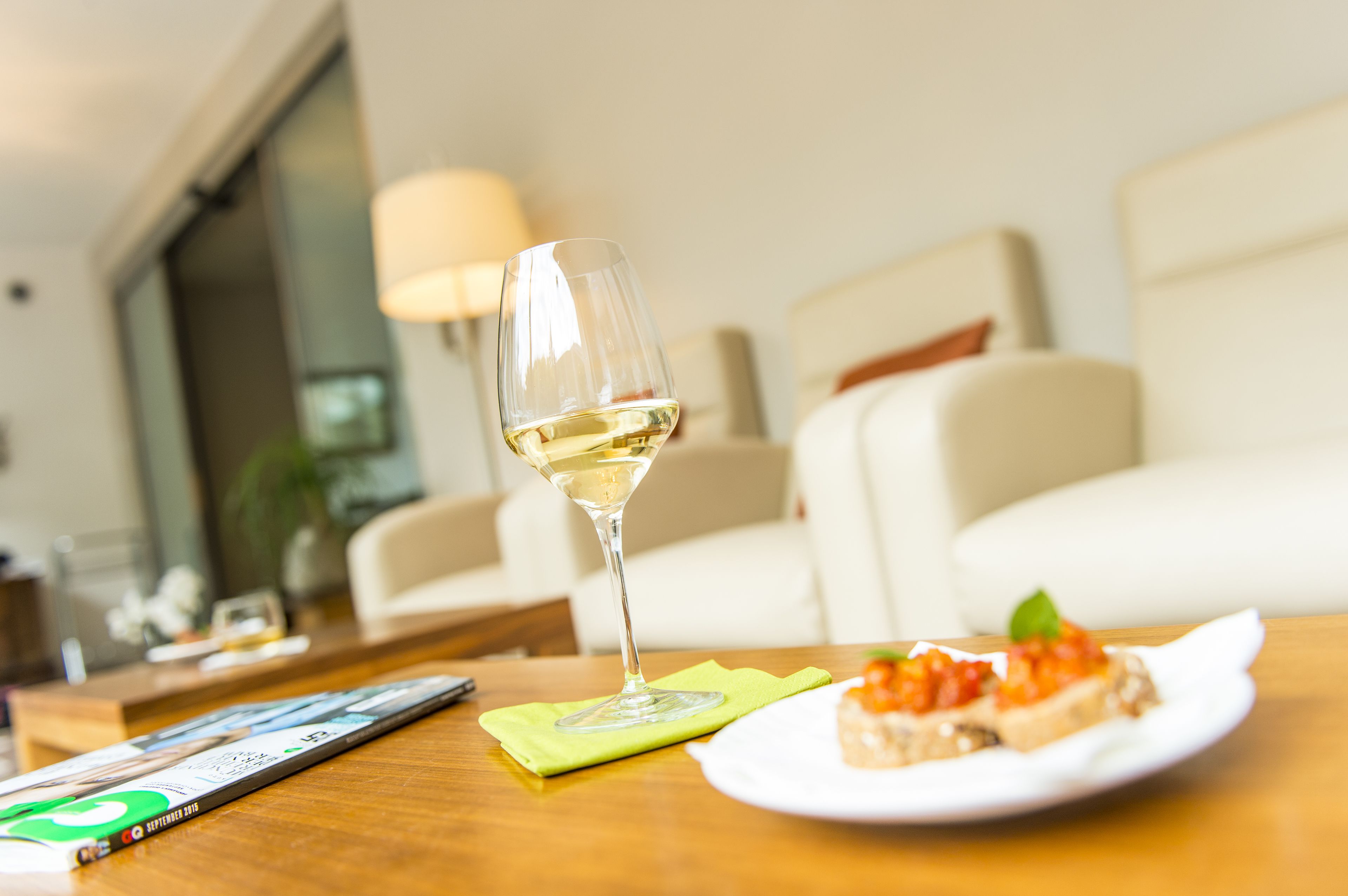 A dining table with a glass of white wine, a plate with a slice of bread and a topping, and a magazine.