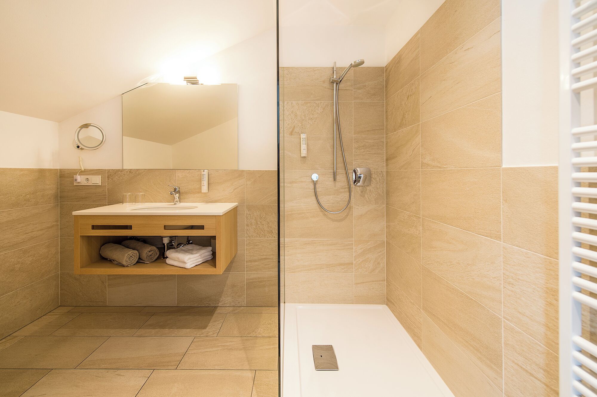 A modern bathroom with a wooden vanity, a glass shower enclosure, and beige tiles.