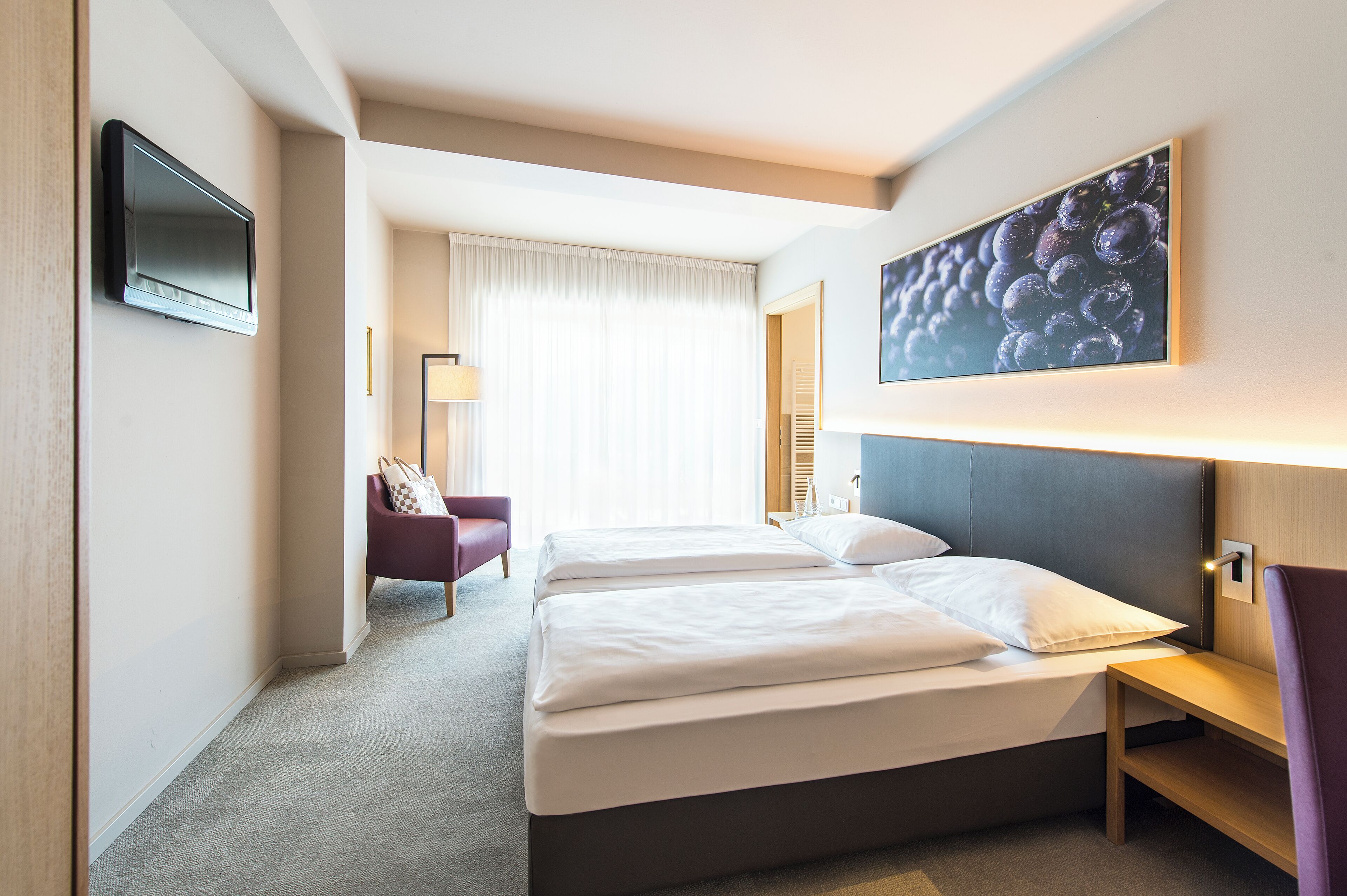 Ein Hotelzimmer mit zwei Betten, einem an der Wand montierten Fernseher, einem Traubenbild und einem Schreibtisch mit Stuhl.