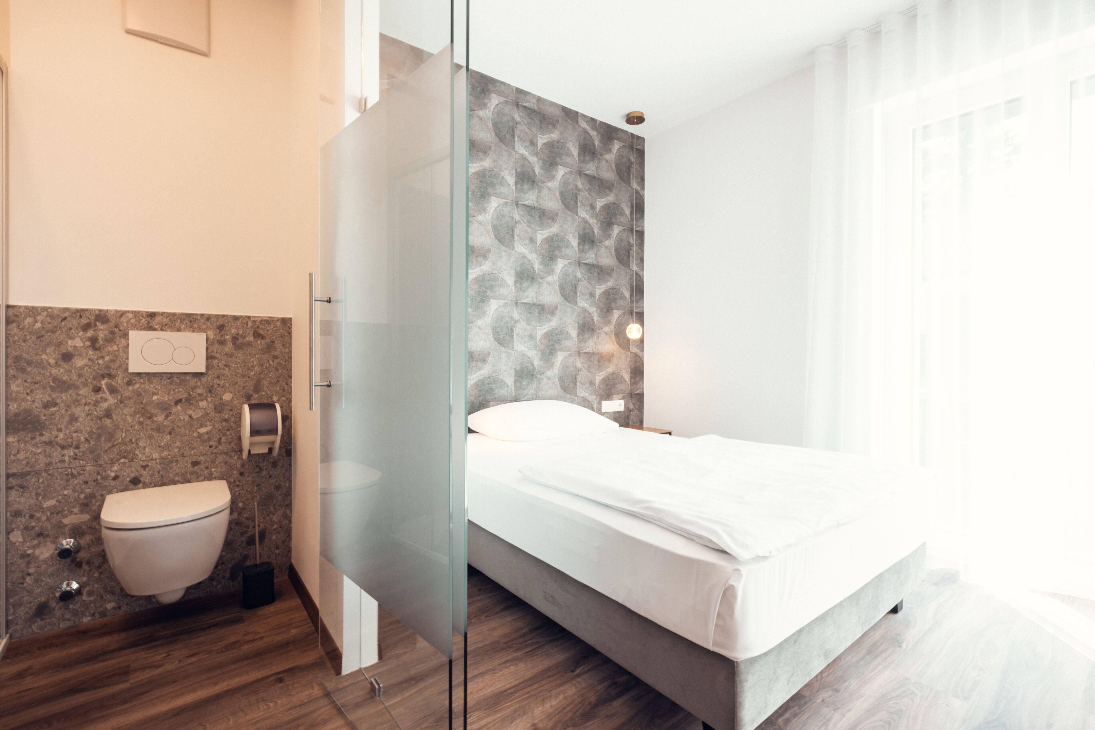 Habitación de hotel moderna con un separador de vidrio que divide el baño del dormitorio, una pared con diseño de mármol, piso de madera y una cama con ropa blanca.