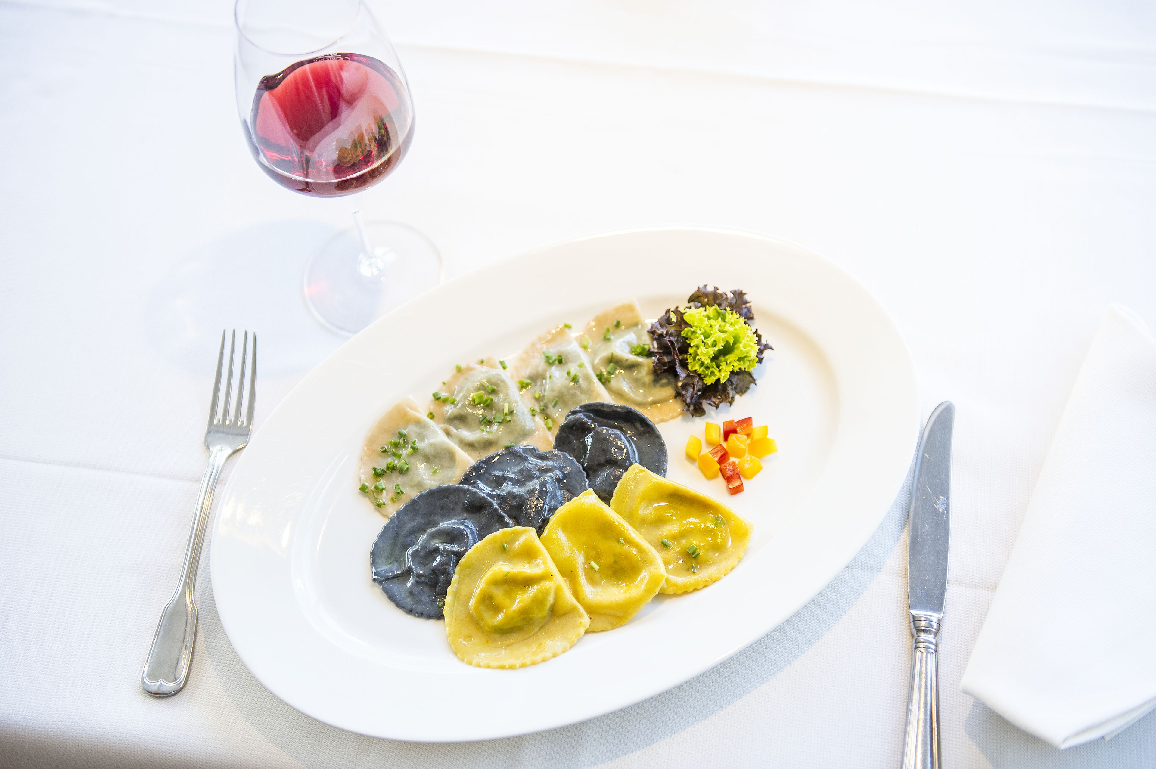 Un plato de ravioles con una ensalada y una copa de vino tinto sobre un mantel blanco.