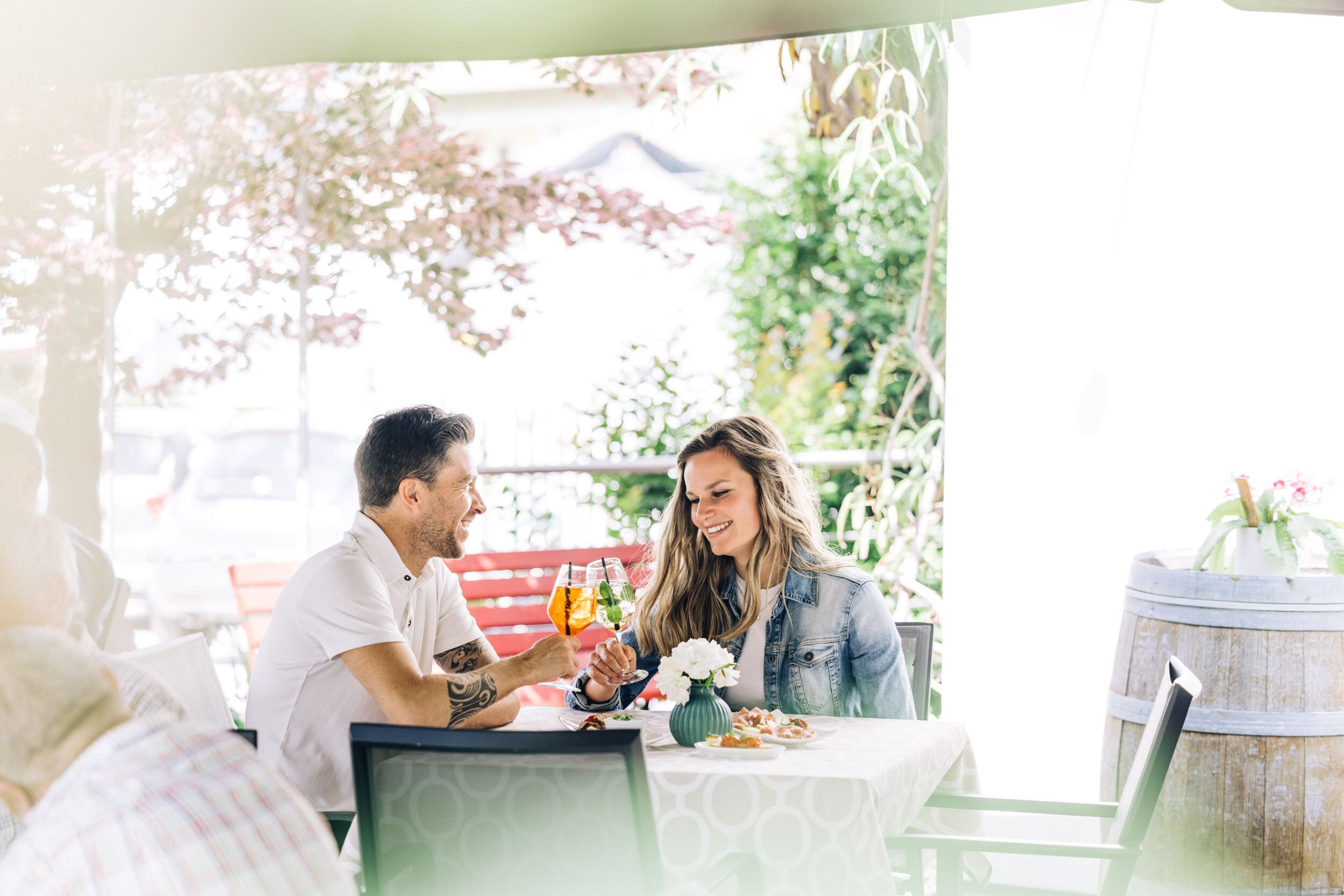 A couple toasting with drinks at an outdoor table with a view of greenery and a wooden barrel.