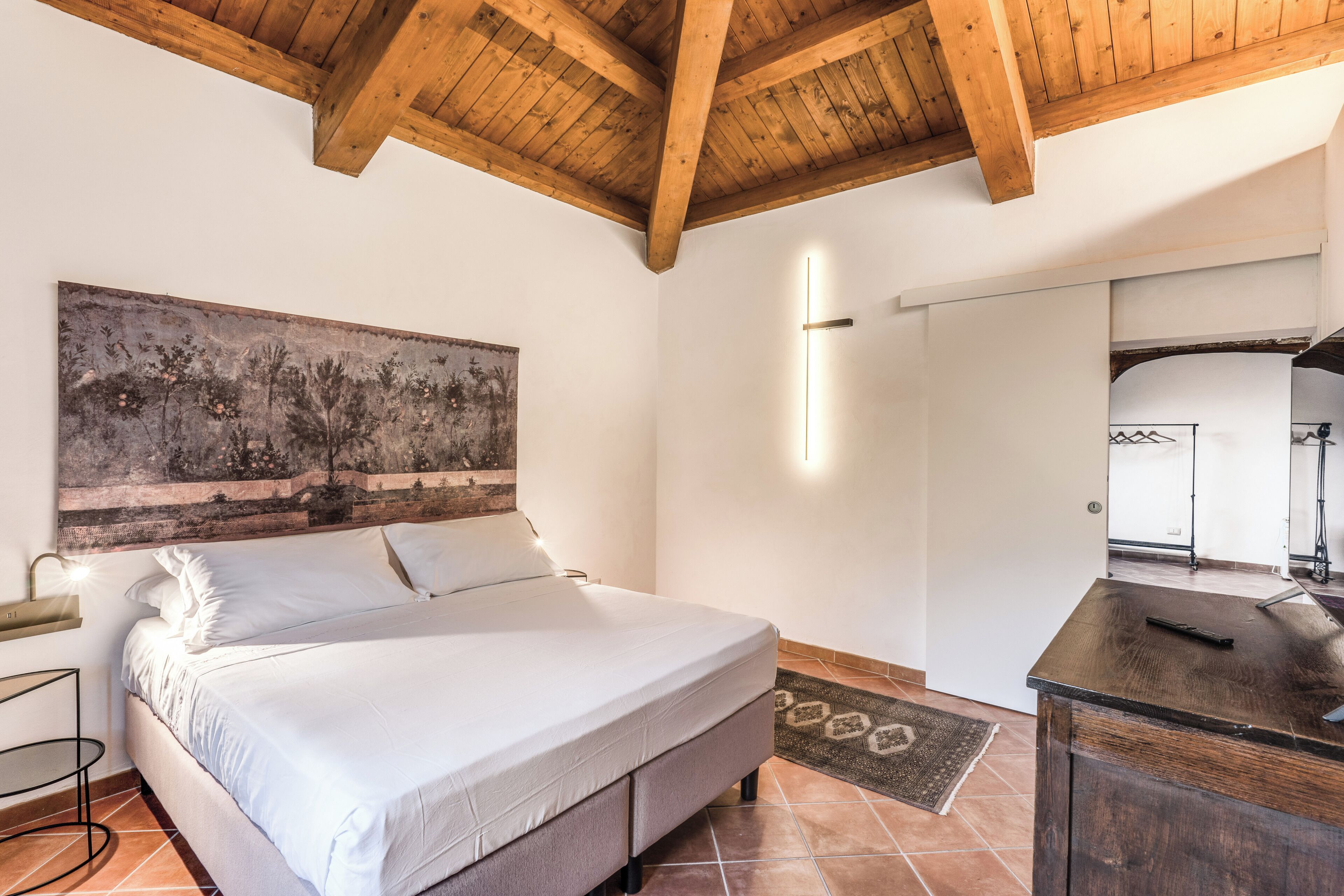 A bedroom with a bed, a large painting, a bedside table, a desk, and a mirror.