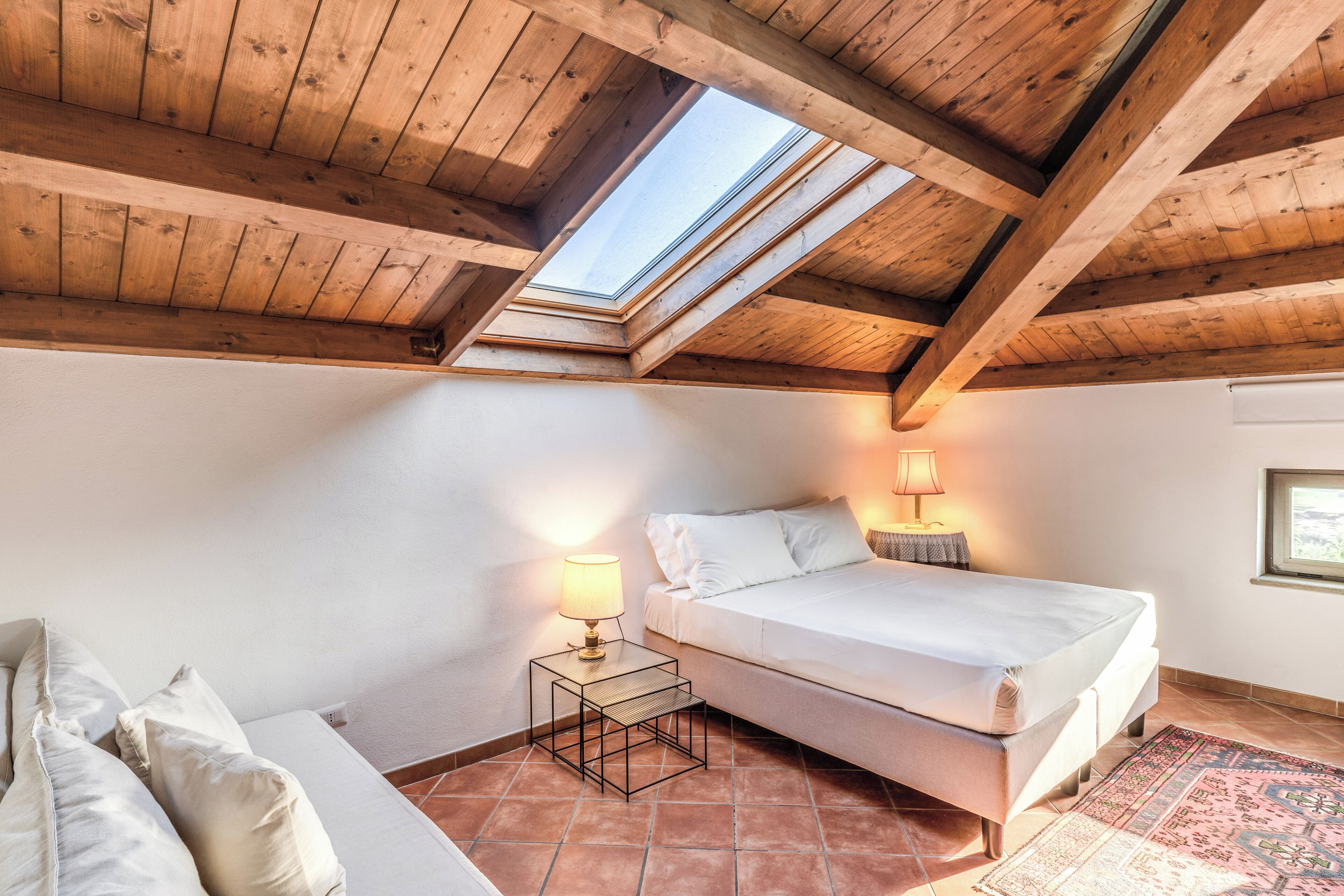 Un dormitorio con tragaluz, una cama, una mesita de noche con lámpara y un sofá.