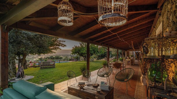 A covered outdoor seating area with a wooden ceiling, wicker chairs, and a view of a grassy landscape.
