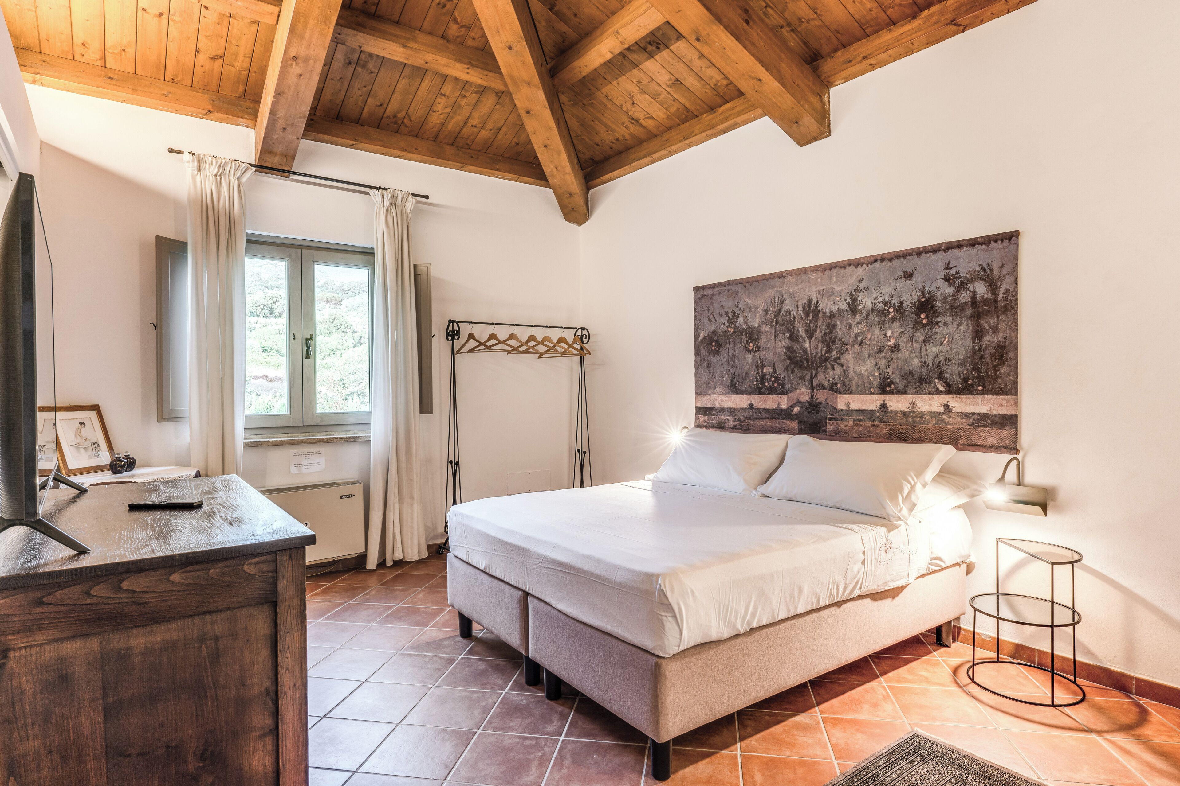 A bedroom with a bed, a desk, a window with curtains, a painting on the wall, and a wooden ceiling.