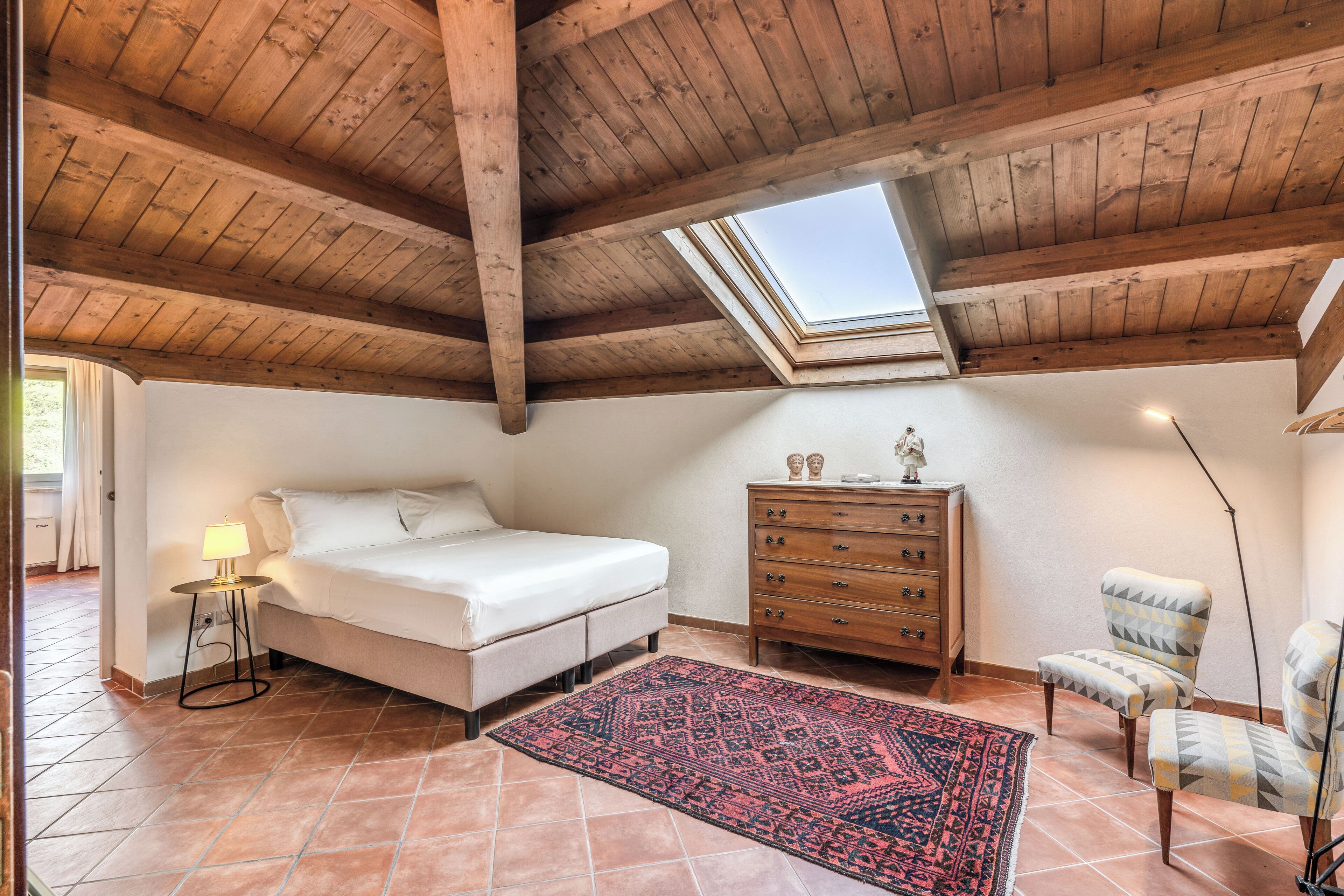 A bedroom with a skylight, a bed, a dresser, a chair, and a patterned rug.