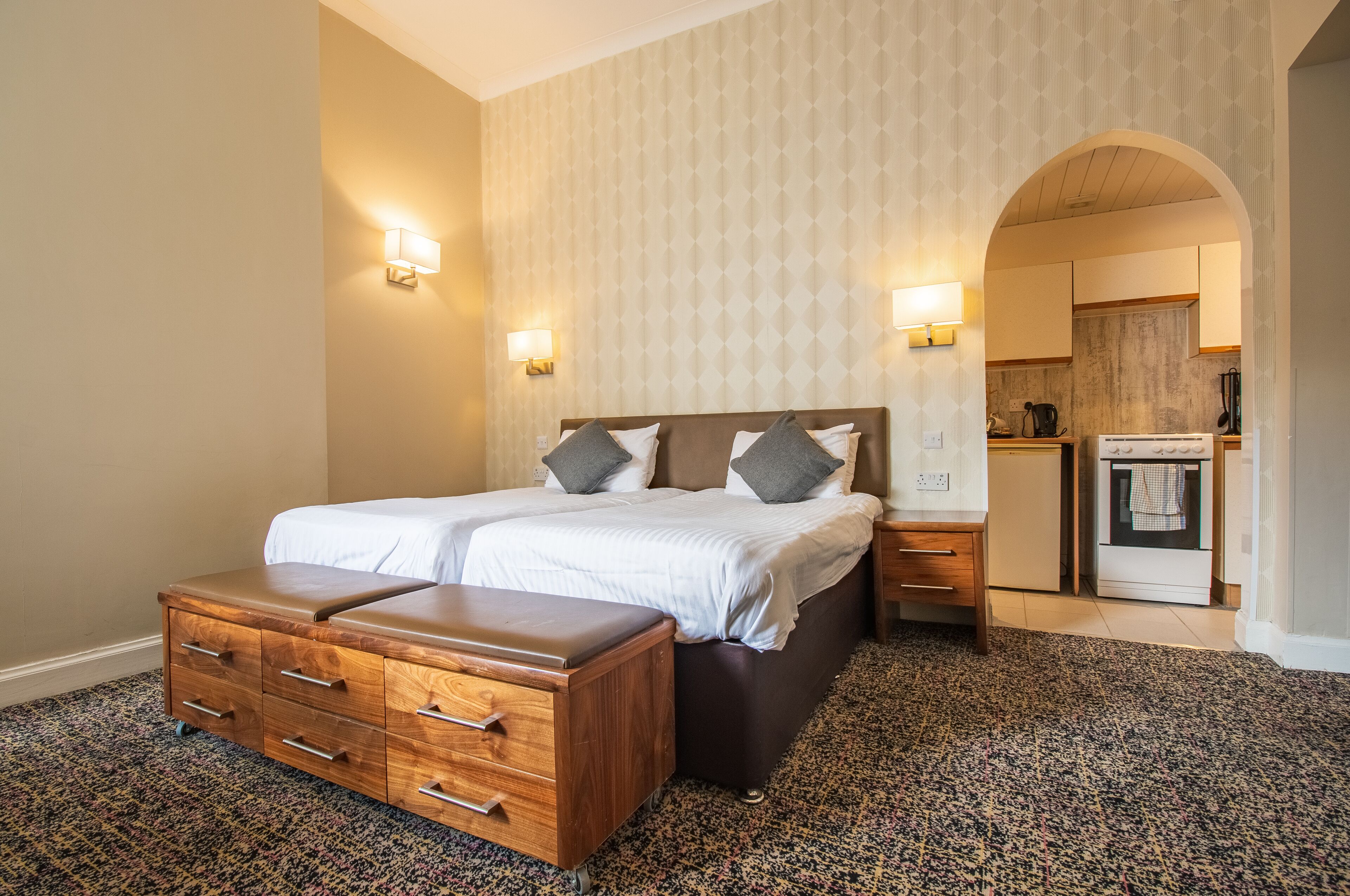 Une chambre avec un lit double, une commode en bois, deux tables de chevet et une cuisine visible à travers une arche.
