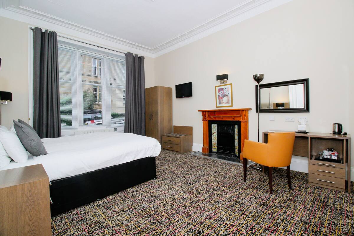 Een hotelkamer met een bed, een bureau, een stoel, een openhaard en een groot raam met gordijnen.