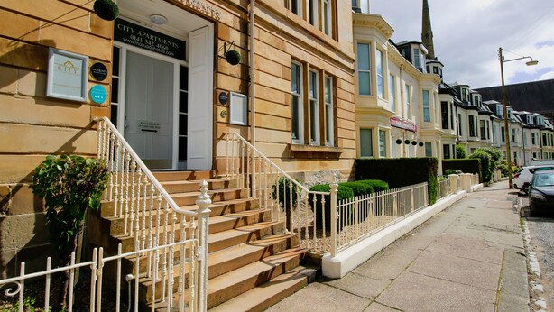 A row of city apartments with a staircase leading to the entrance, a signboard, and a neighboring church spire. - City Apartments Glasgow (Glasgow)