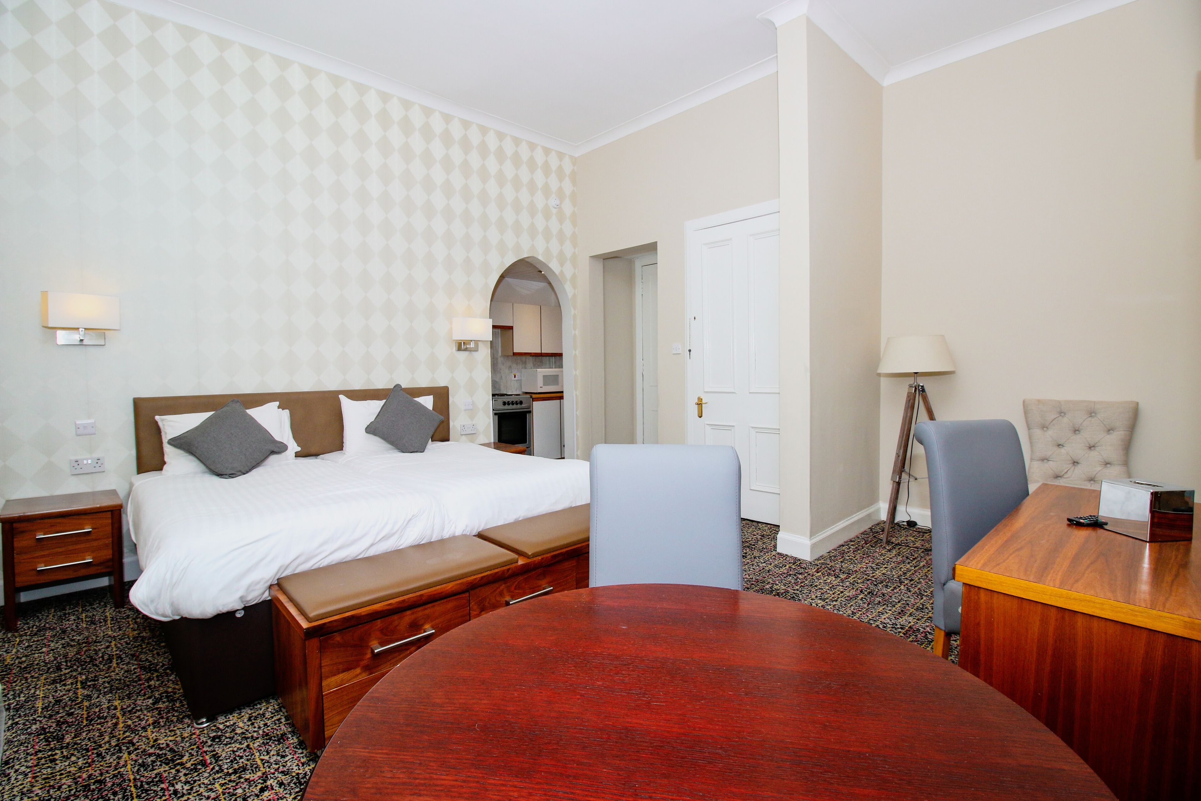 Una habitación de hotel con cama, mesita de noche, lámpara, silla y una mesa de comedor de madera.