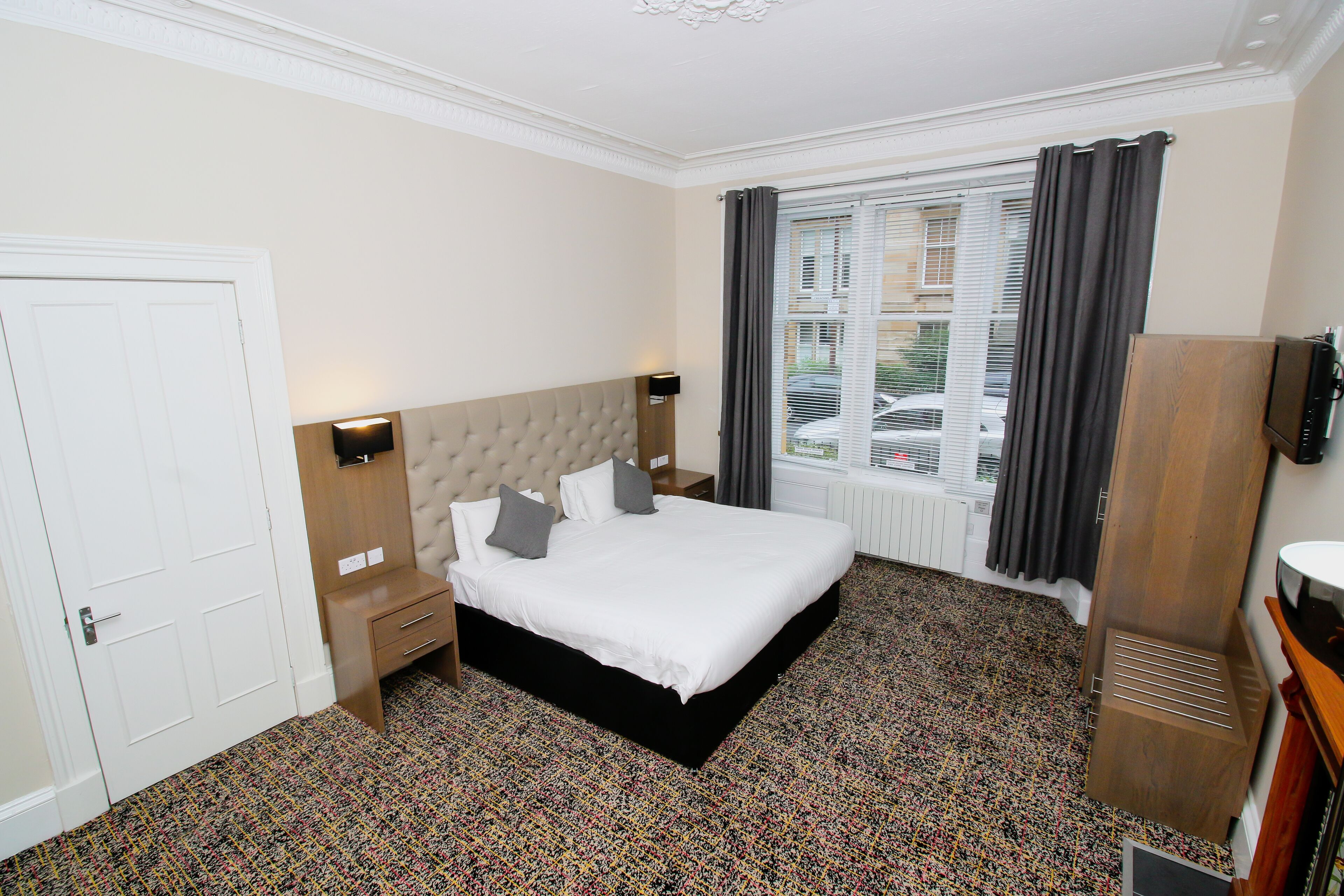 Habitación de hotel con cama, mesita de noche, cómoda, televisor y ventana con cortinas.