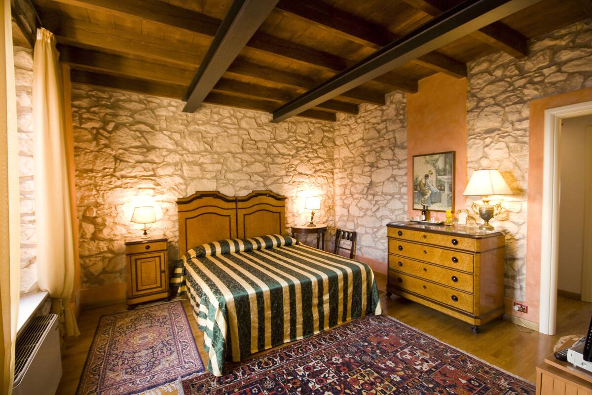 A bedroom with a stone wall, wooden beams, a bed with a striped bedspread, two bedside tables with lamps, a dresser, and a rug.