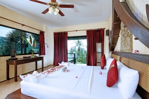 A hotel room with a large bed, red curtains, a wooden headboard, a ceiling fan, and a view of palm trees. - Boomerang Village Resort Kata (Karon)