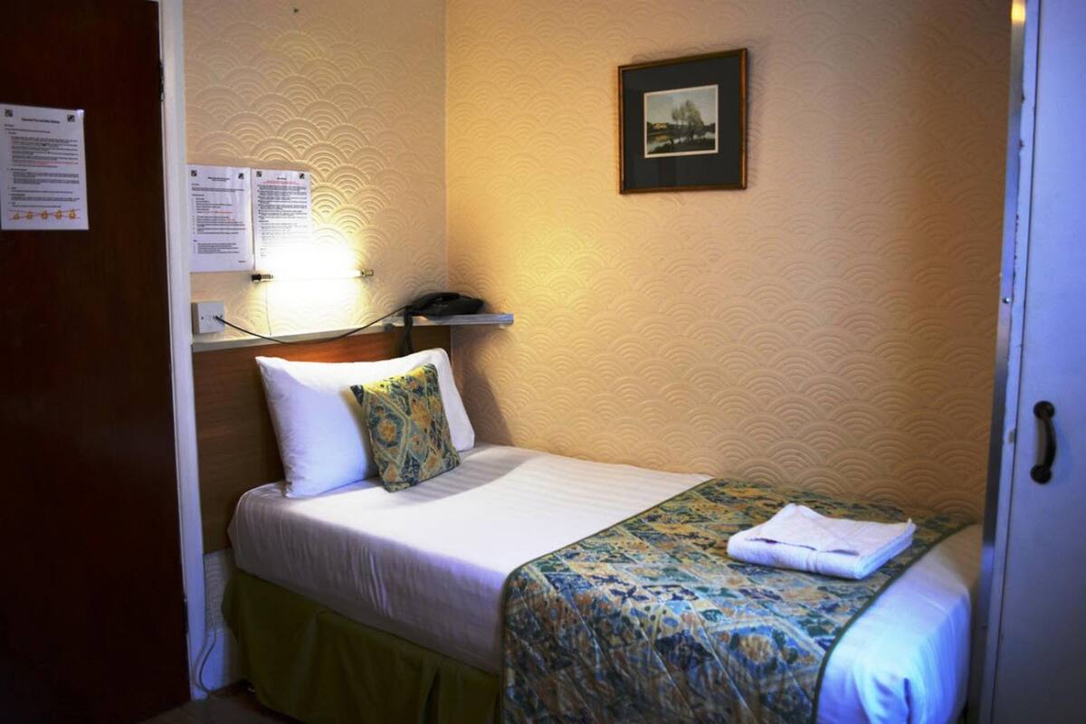 A hotel room with a bed, a bedside table, a lamp, a framed picture on the wall, and a door.