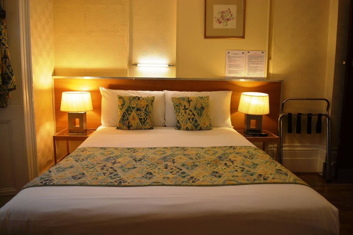 A hotel room with a bed, two bedside lamps, a nightstand, a chair, and a wall-mounted radiator.