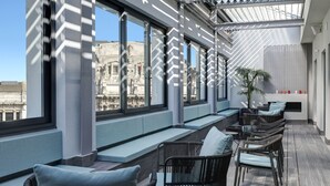 Terraza en la azotea con mobiliario, ventanas amplias y vistas a un edificio histórico.